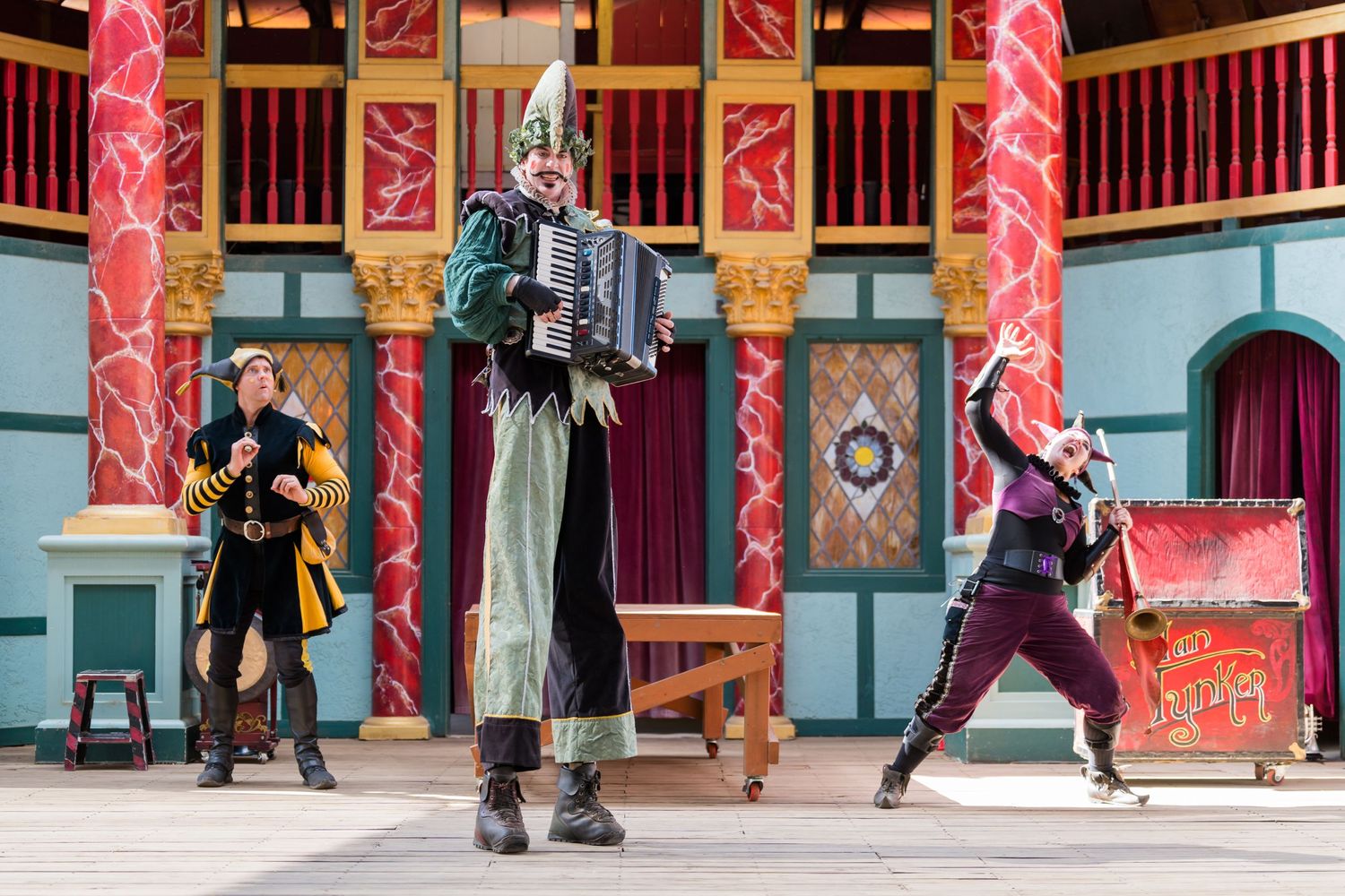 Performers in medieval costumes entertain on a decorated stage with colorful columns and backdrops.