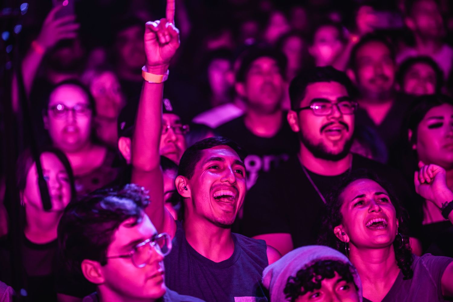 Enthusiastic concert crowd illuminated in purple light cheering and singing along at a live music performance.