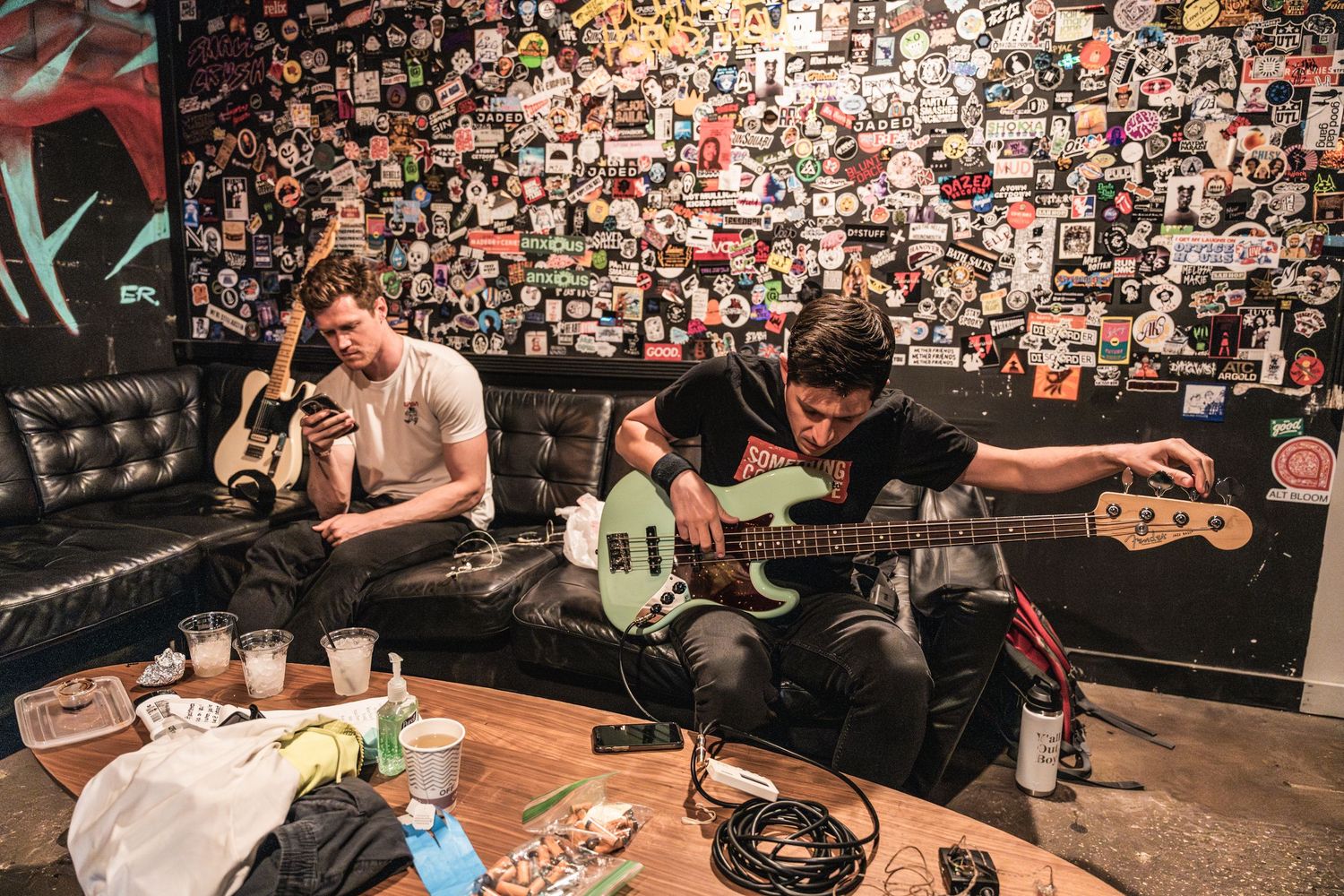 Band members relax in front of a wall covered in photos during a casual backstage moment with drinks and equipment.