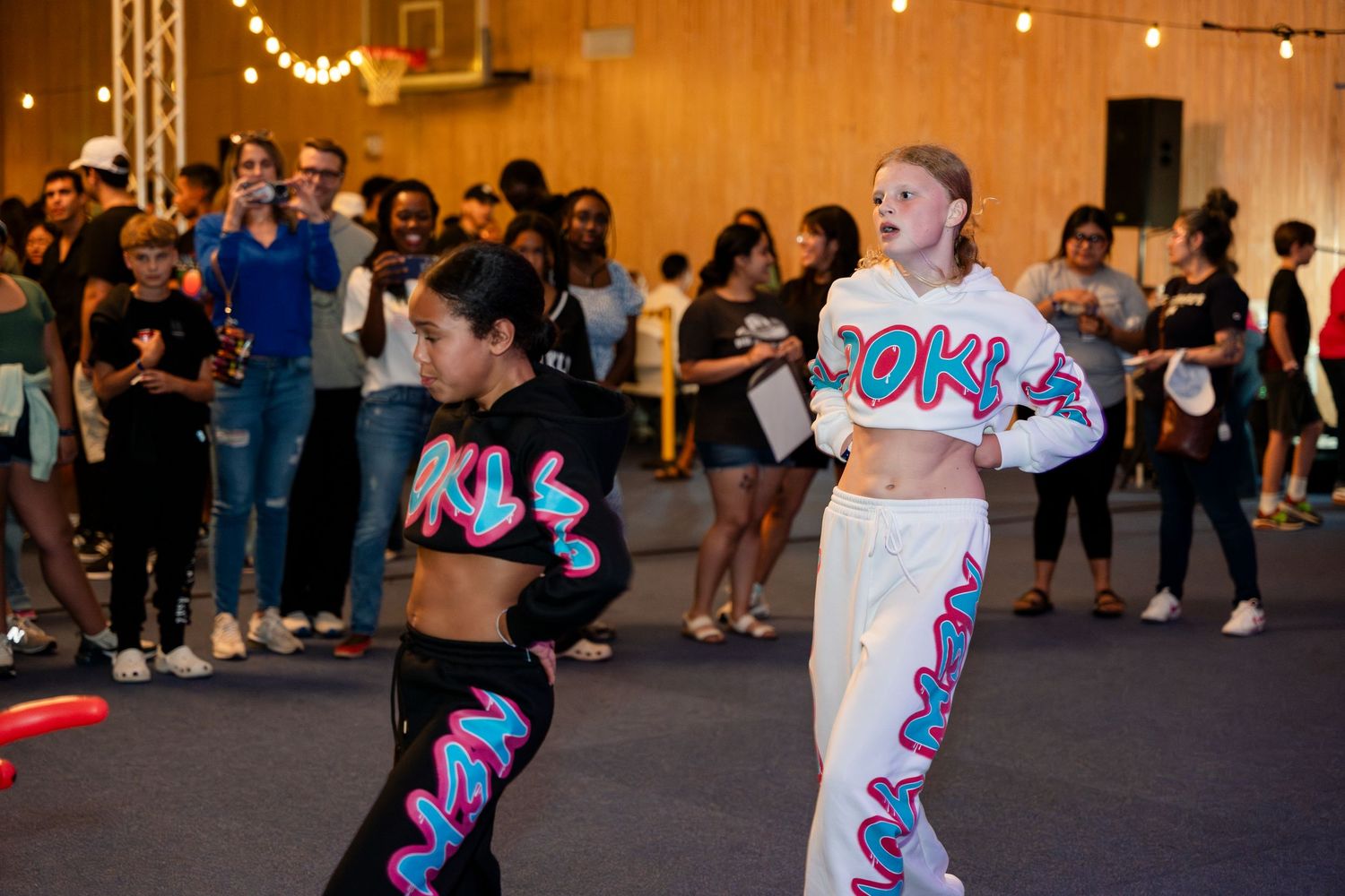 Two dancers perform energetically in white and black outfits with colorful designs as a crowd watches in the background.