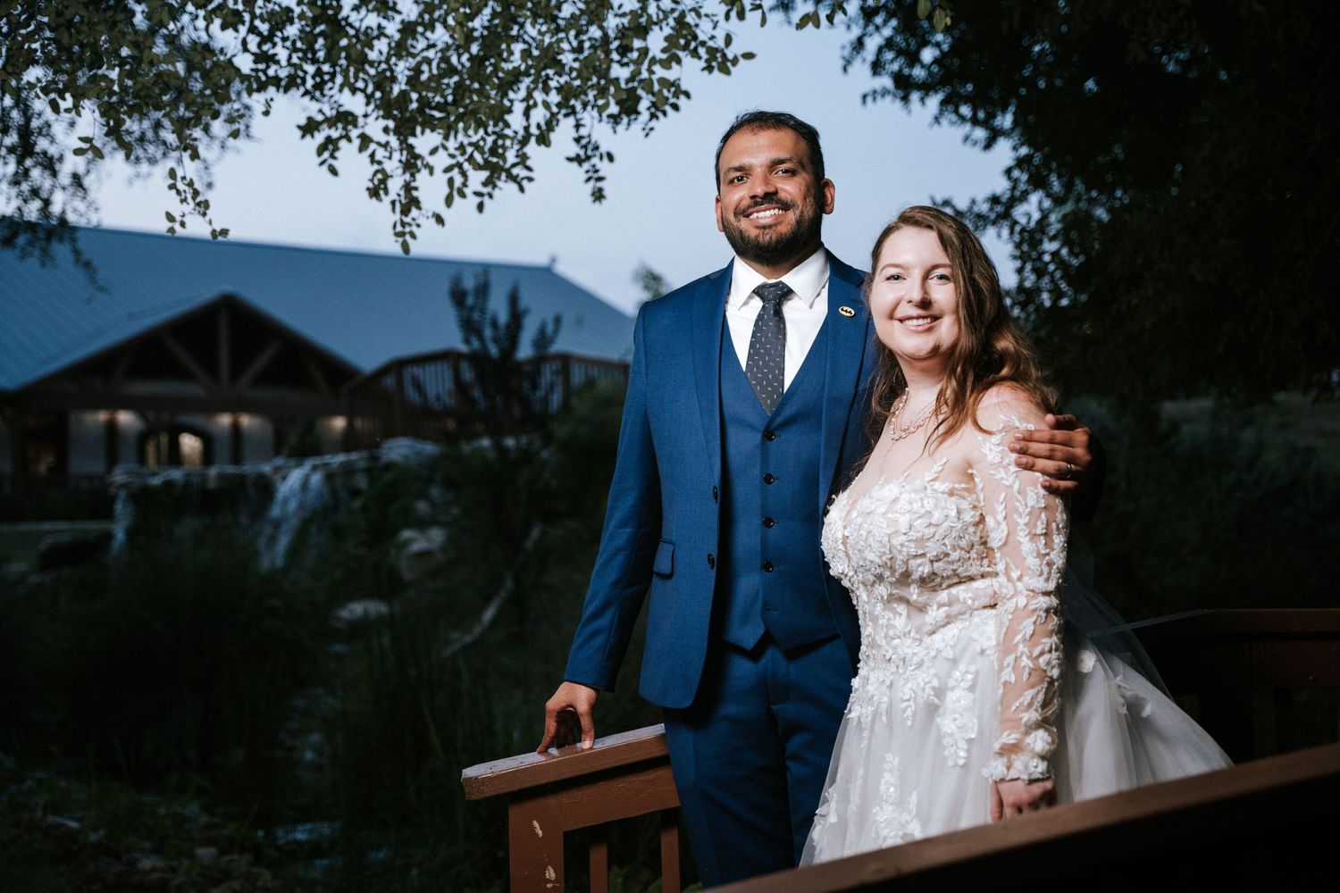 A couple poses outdoors for a portrait with mountains and a rustic building visible in the background.