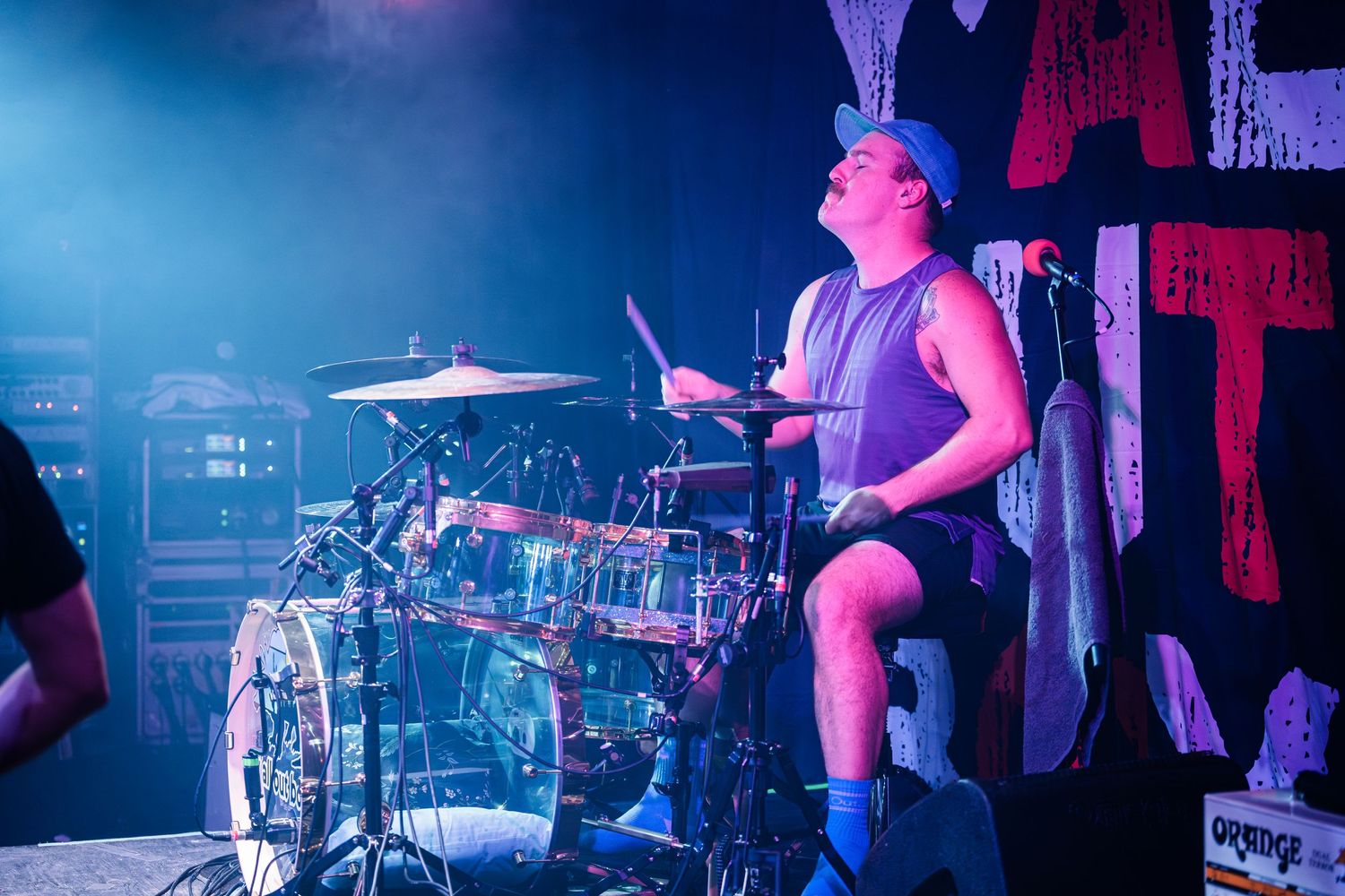 An energetic drummer performs on stage bathed in vibrant blue lighting during a live concert performance.