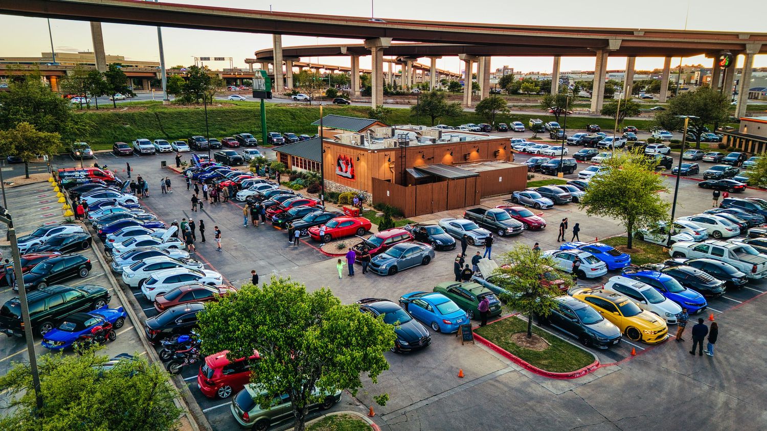 Aerial view of a crowded parking lot at a car meet with colorful vehicles surrounding a brick building.