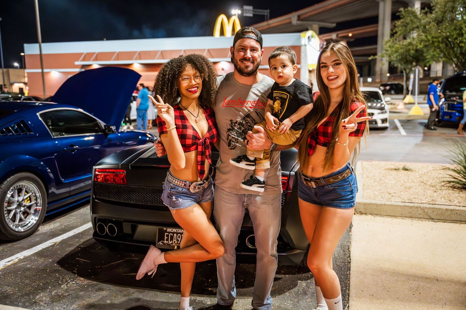 Group poses together with small dog at car meetup outside McDonald's at night.