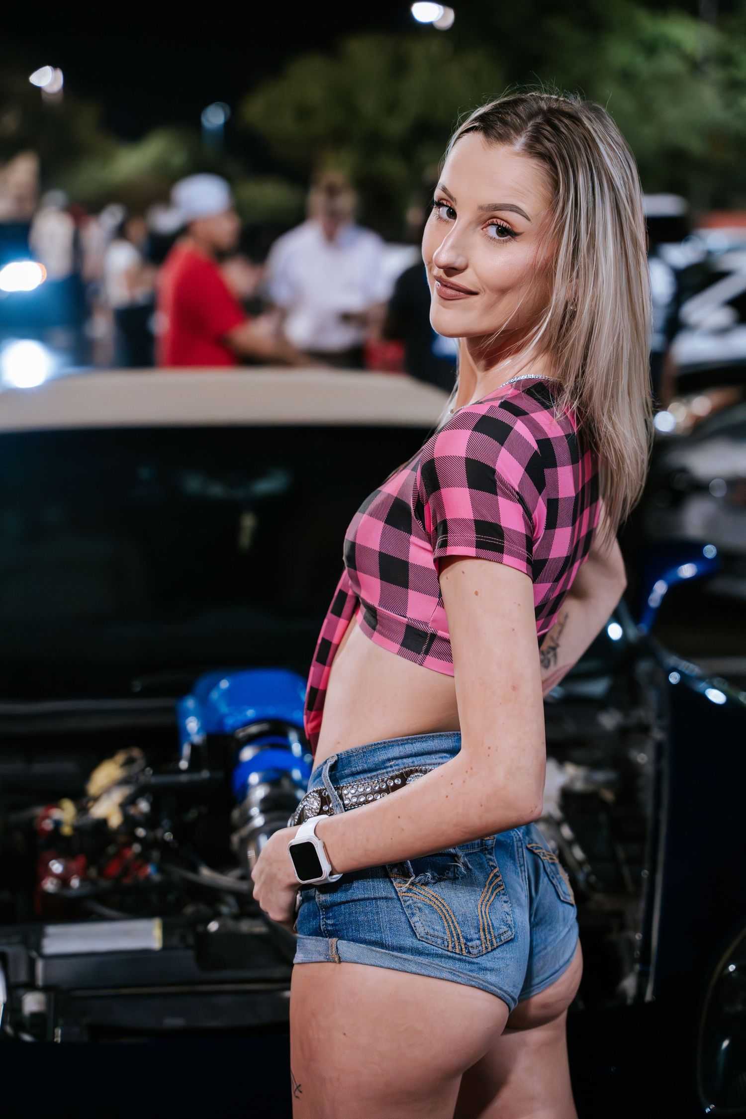 Young car enthusiast in pink plaid crop top and shorts poses beside performance vehicles at a car meet.