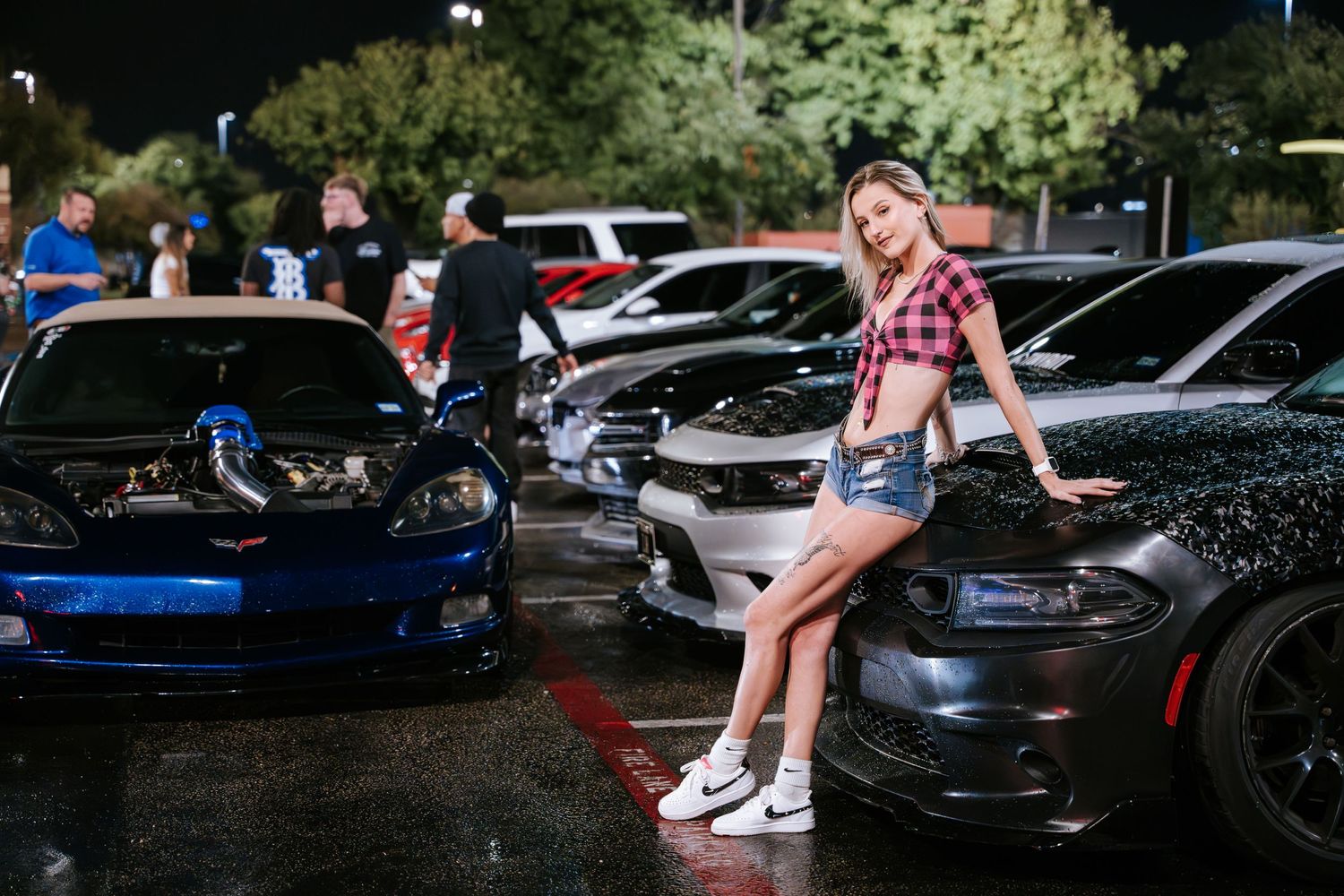 Person sitting on hood of dark vehicle at nighttime car show.