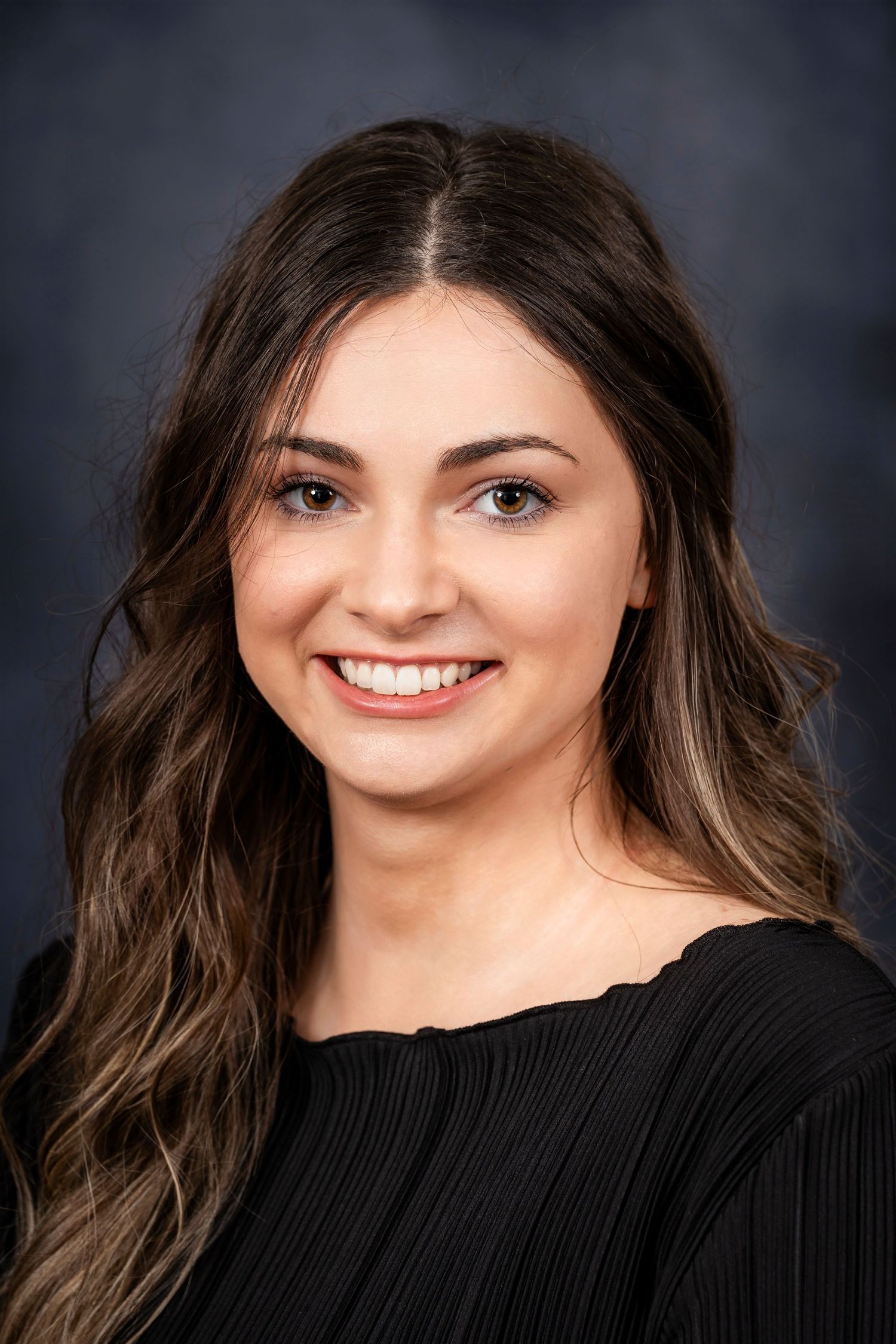 Professional portrait of a person with long wavy brown hair wearing a black pleated top.