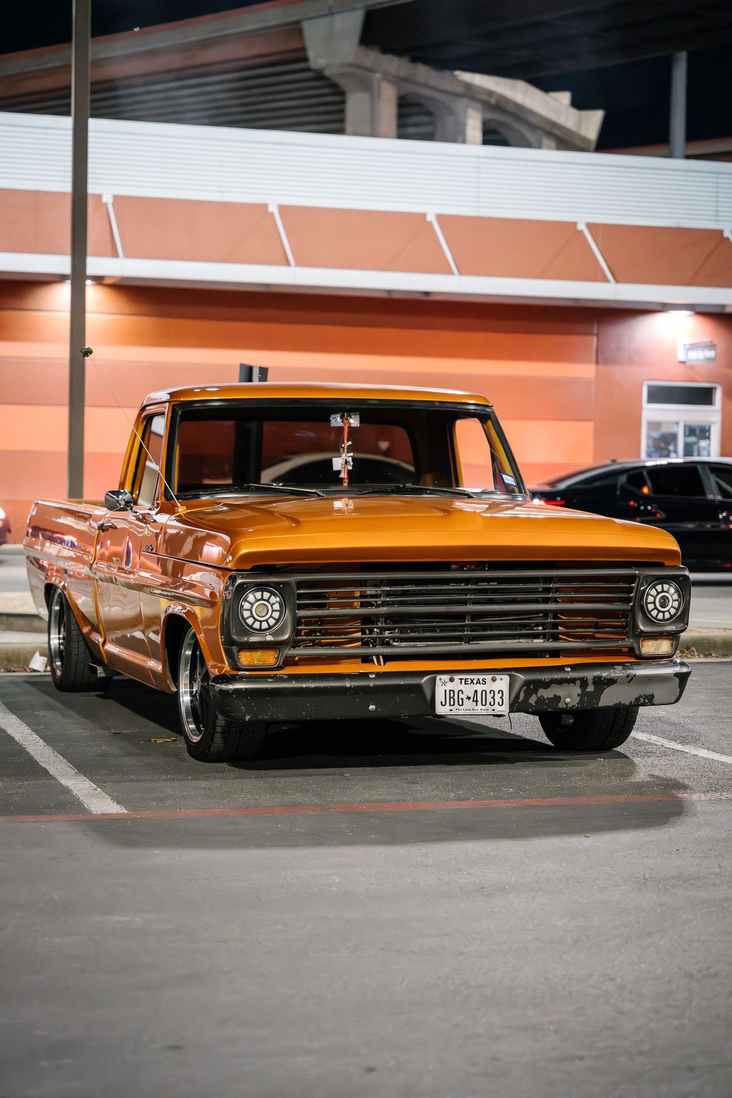 Restored vintage Ford F-100 pickup in orange metallic paint parked in retail lot at night.
