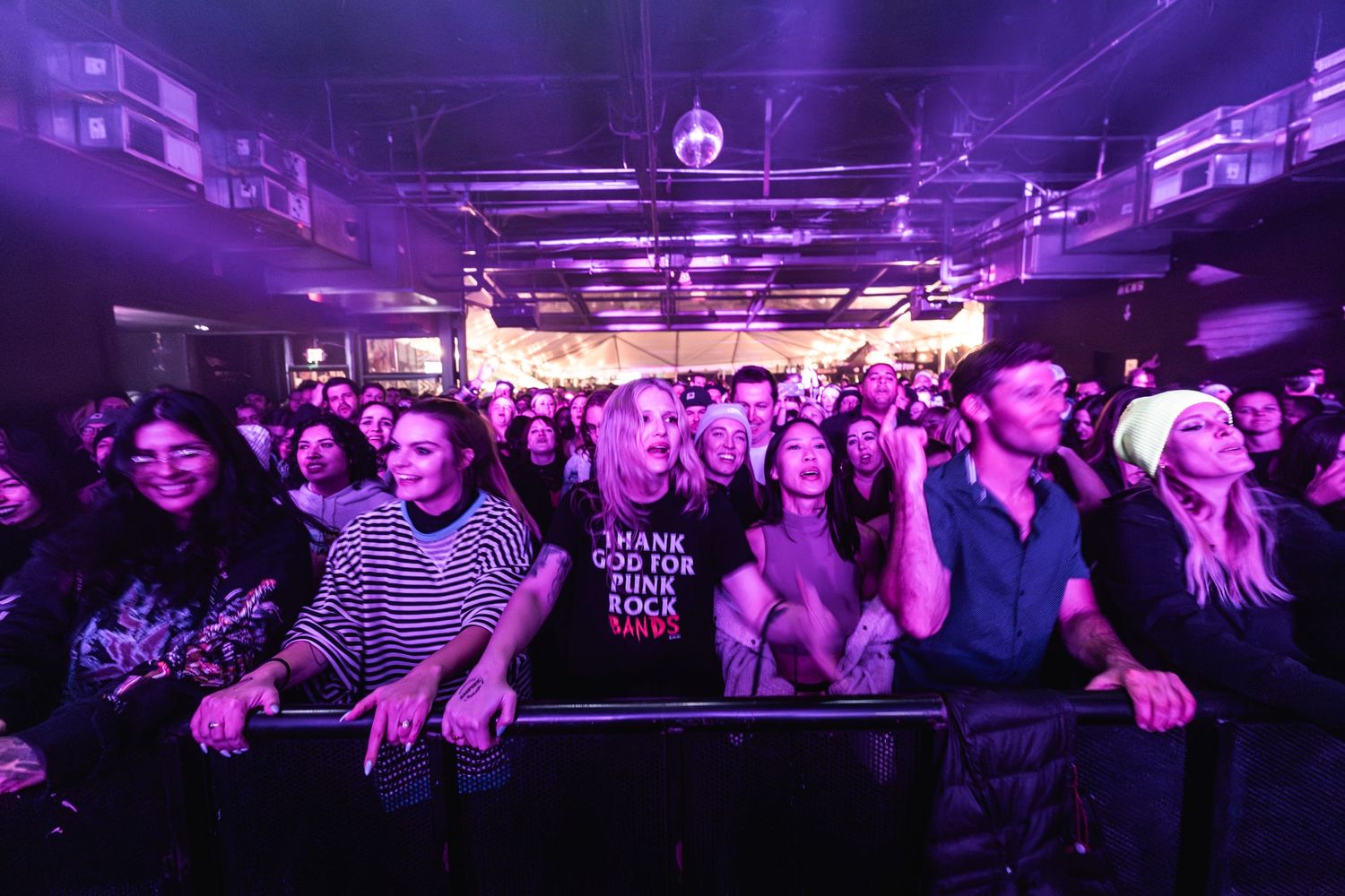 Large audience enthusiastically watching performance in purple-lit concert venue.