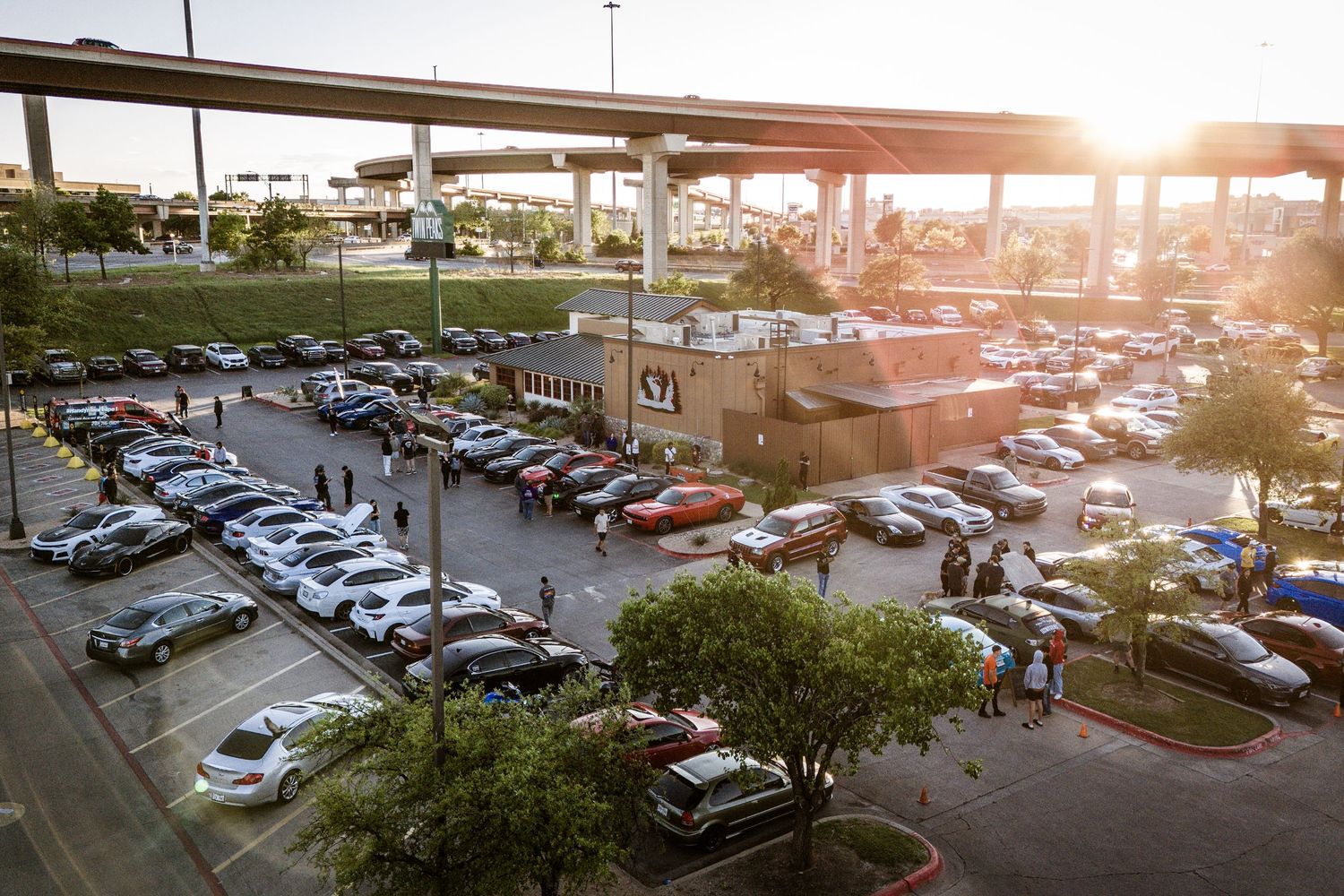Bird's eye view of a packed parking lot during golden hour with highway overpass visible.