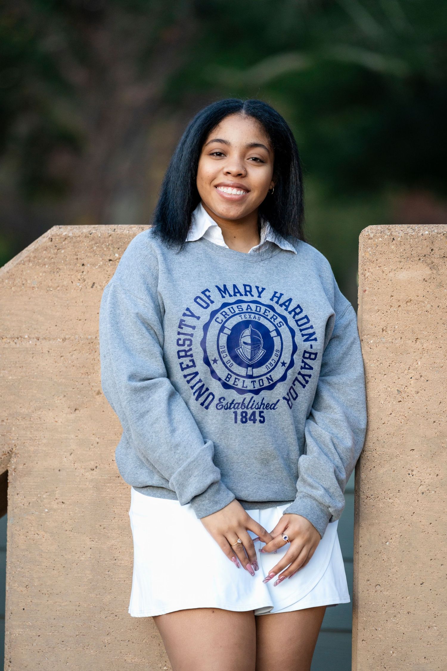 A graduate in grey University of Mary Hardin-Baylor sweatshirt poses outdoors.