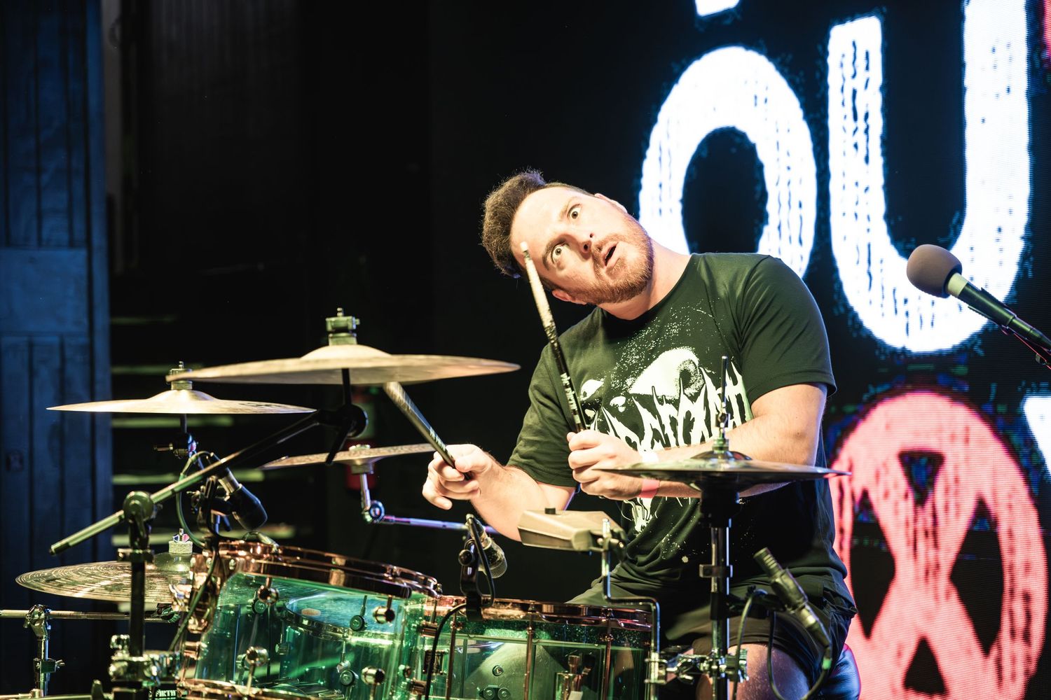 Drummer performs energetically behind drum kit on stage with illuminated backdrop.