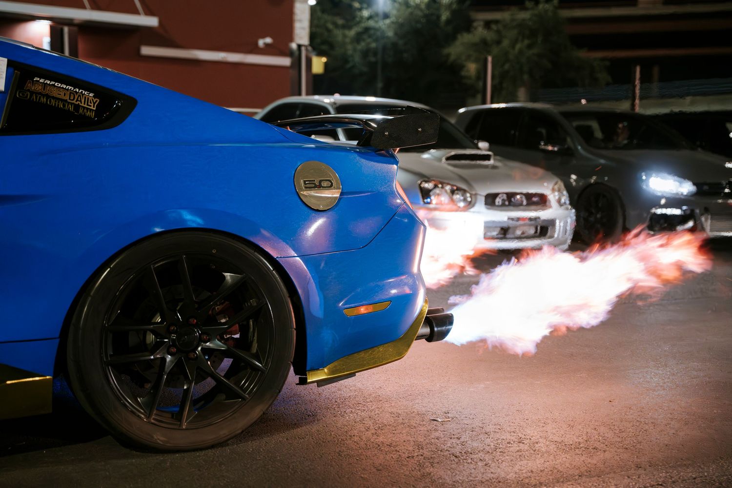 Blue sports car shooting flames from exhaust in dark parking lot.