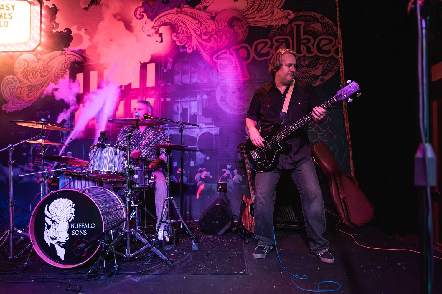 Bass guitarist performs on stage with purple lighting and drum set in background.