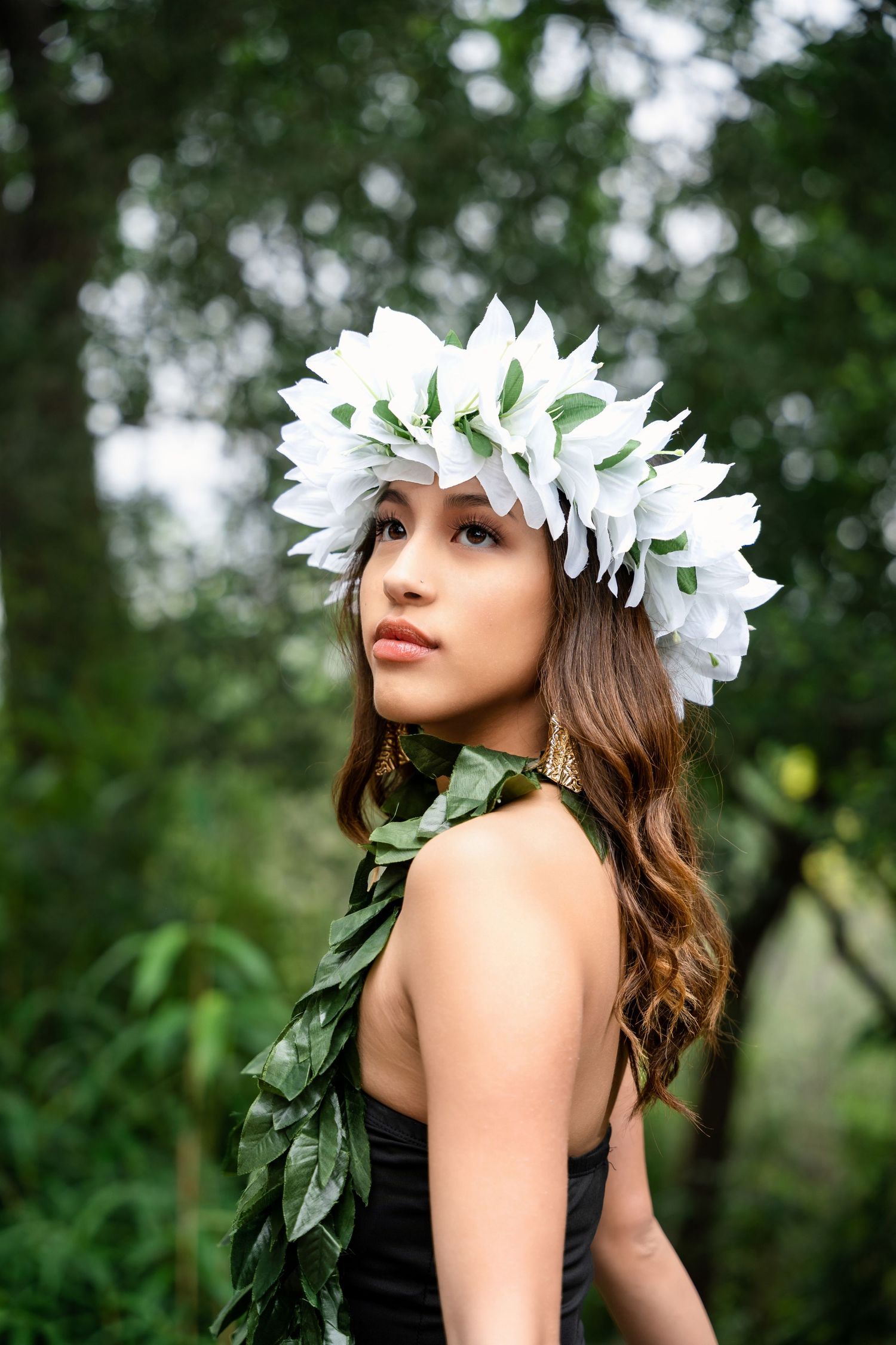 Side profile portrait featuring white flower crown and green leaf garland against forest backdrop.