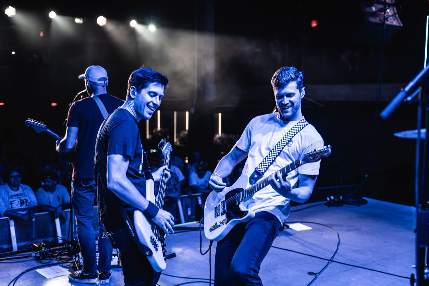 Band members performing energetically with guitars during concert with blue lighting effects.