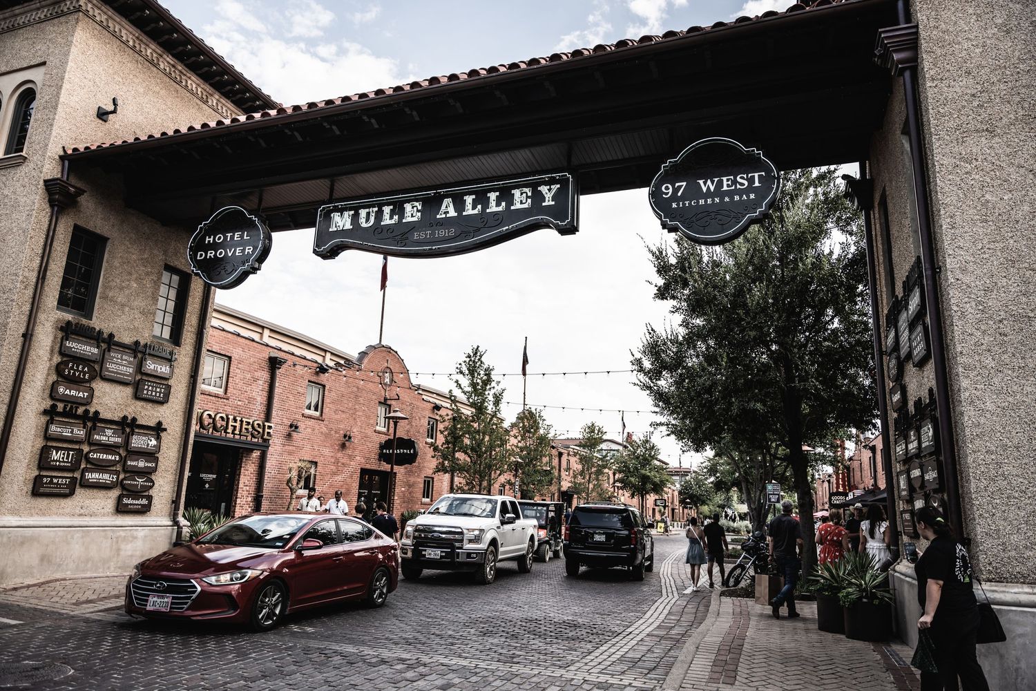 A rustic brick-lined alley entrance with 'Mule Alley' signage spanning across the top and cars parked underneath.