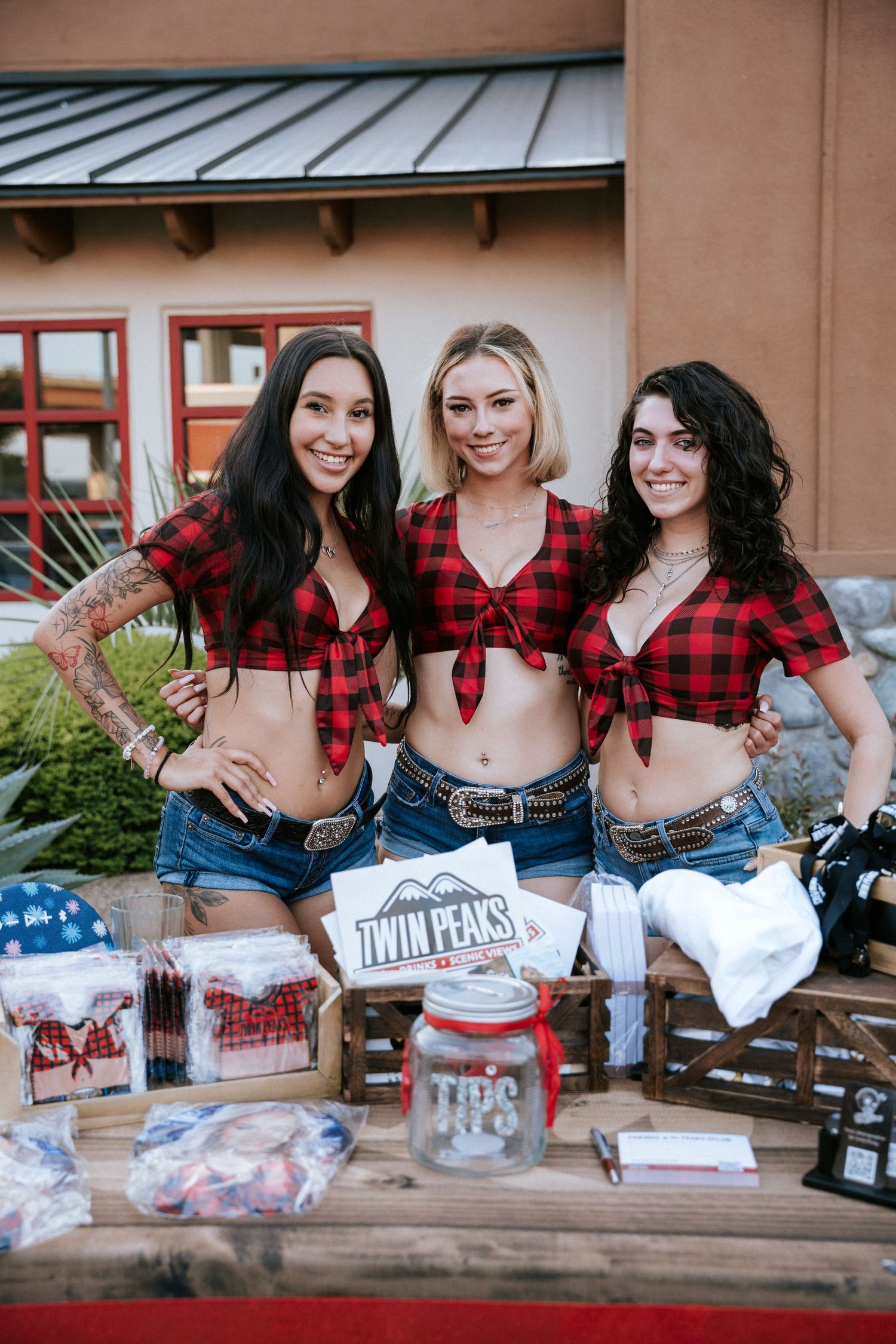 Three women in plaid crop tops and denim shorts selling merchandise at an outdoor vendor table.