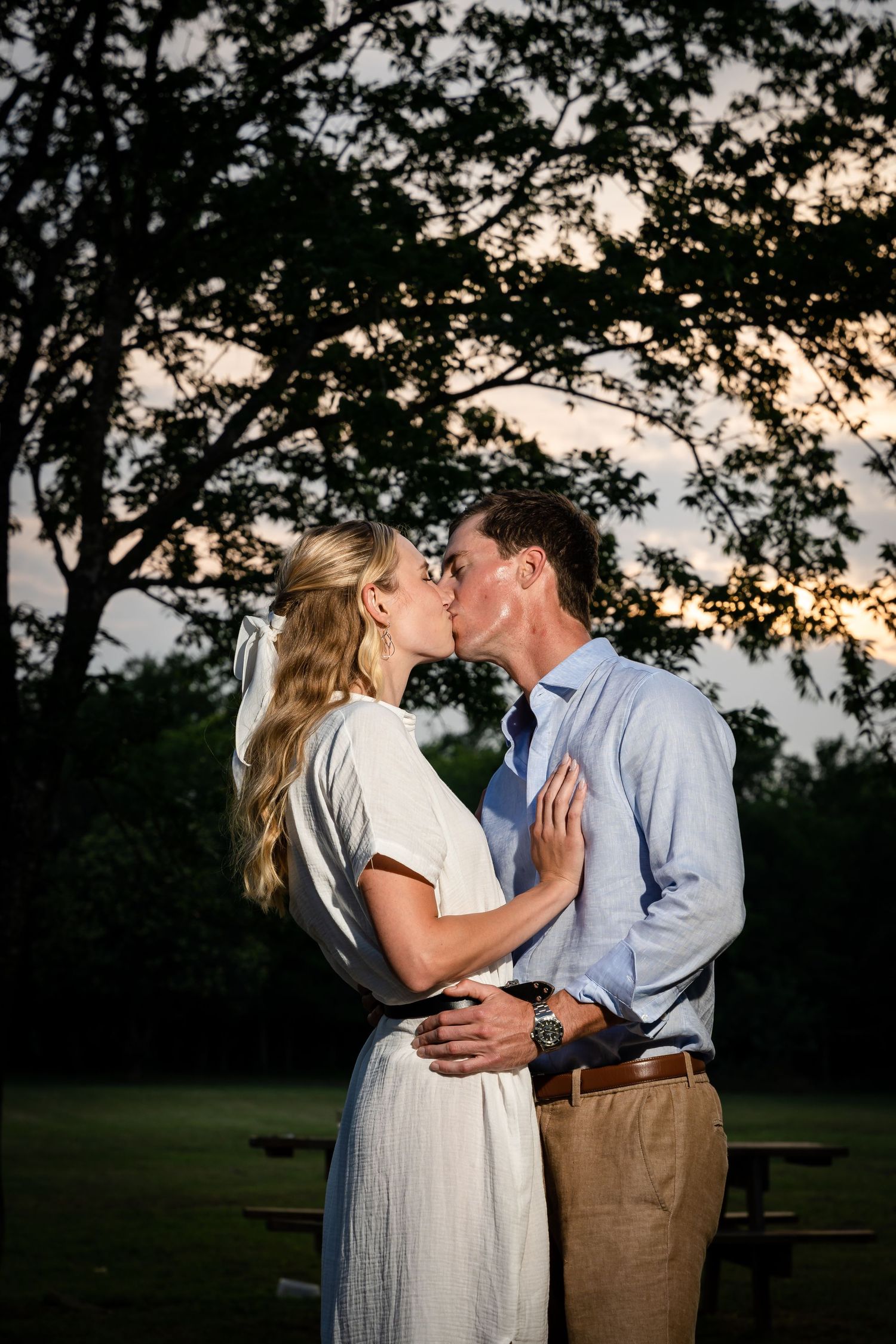 Intimate couple shares a kiss against a dramatic sunset sky and tree silhouettes.