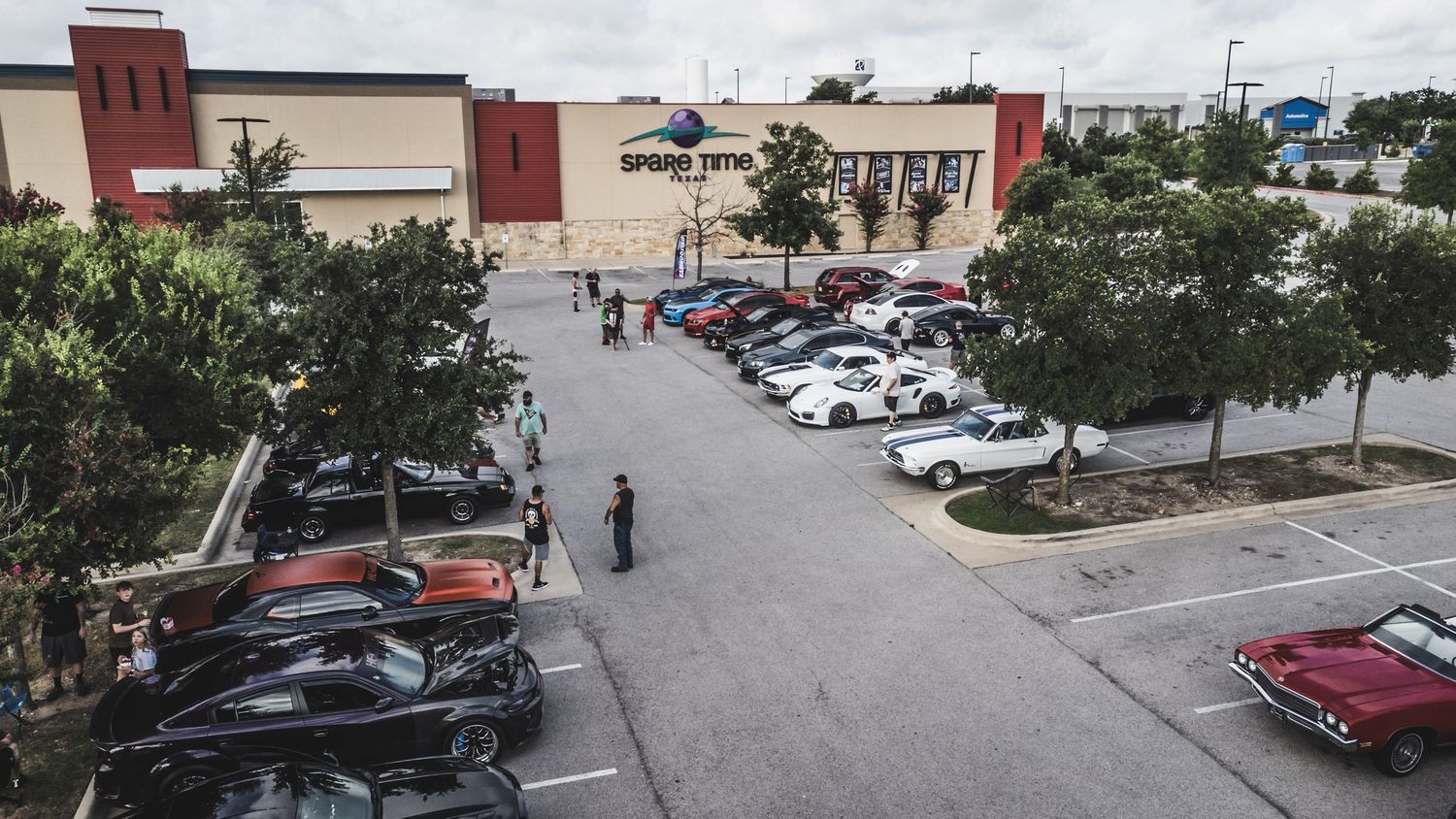 Overhead view of cars parked outside a commercial building during an auto meet event.