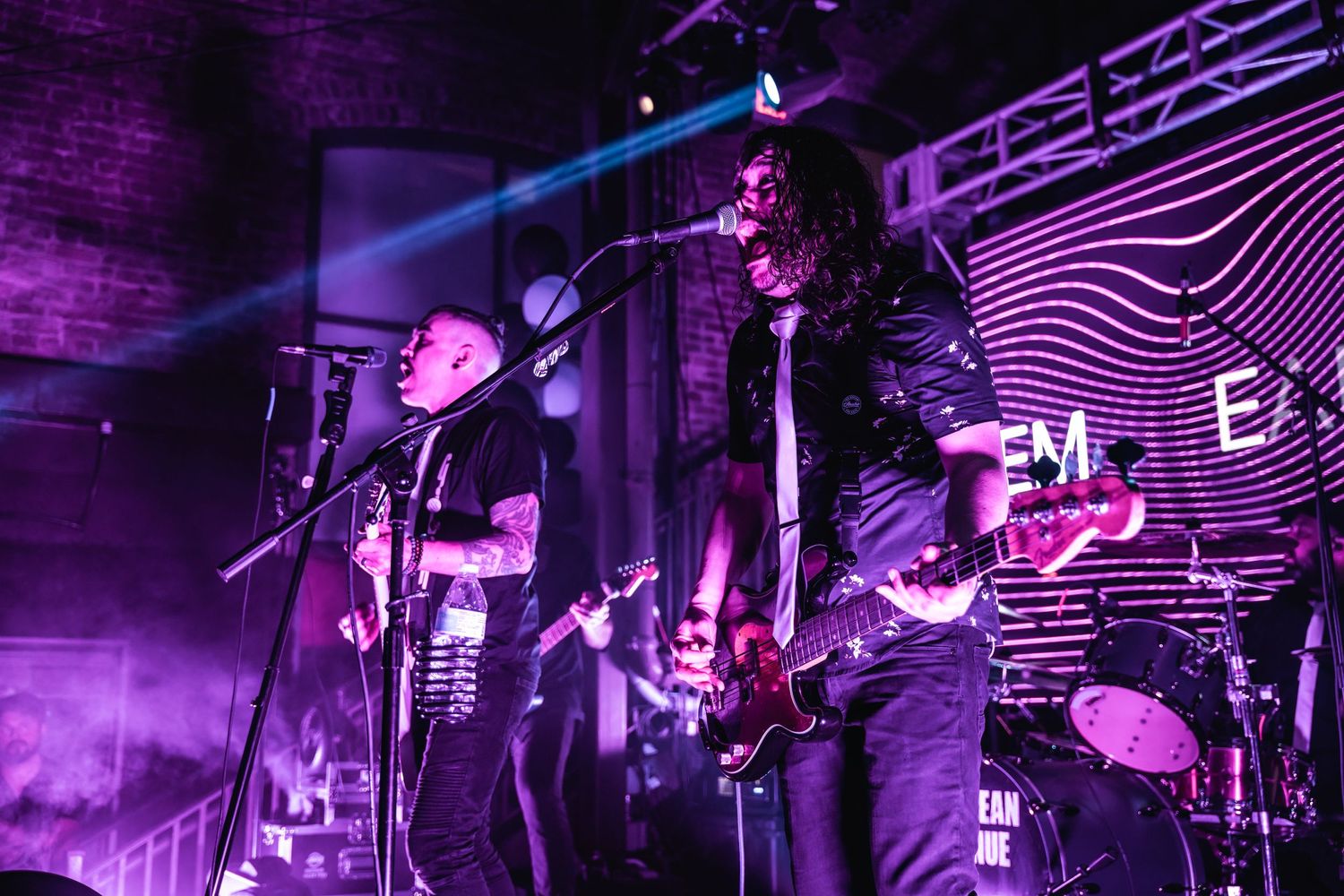 Band performing on stage with purple lighting and wavy backdrop design.