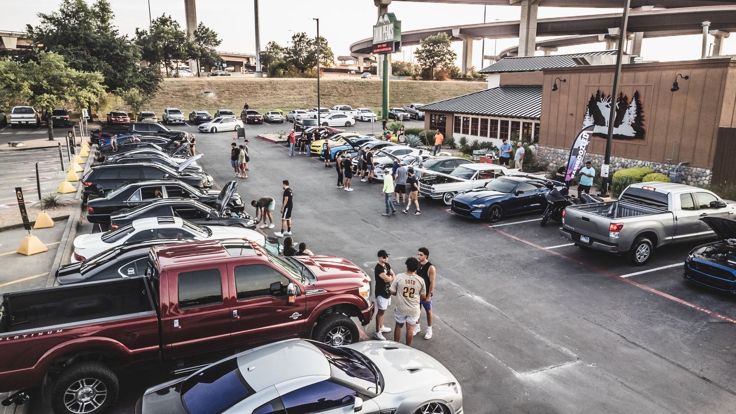 Aerial view of outdoor car meet showing various vehicles parked in commercial lot.