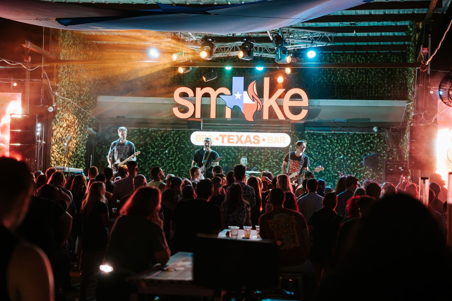 Illuminated 'Smoke' sign glows above concert crowd with colorful stage lighting and live band performing.