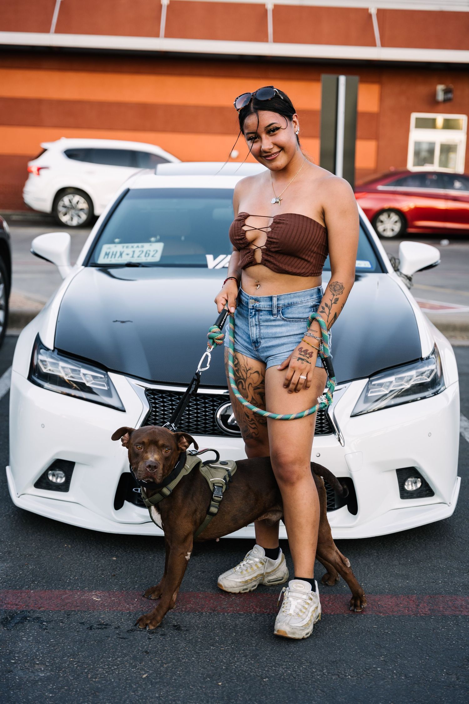A person poses with their brown pit bull mix dog in front of a white Lexus sedan.