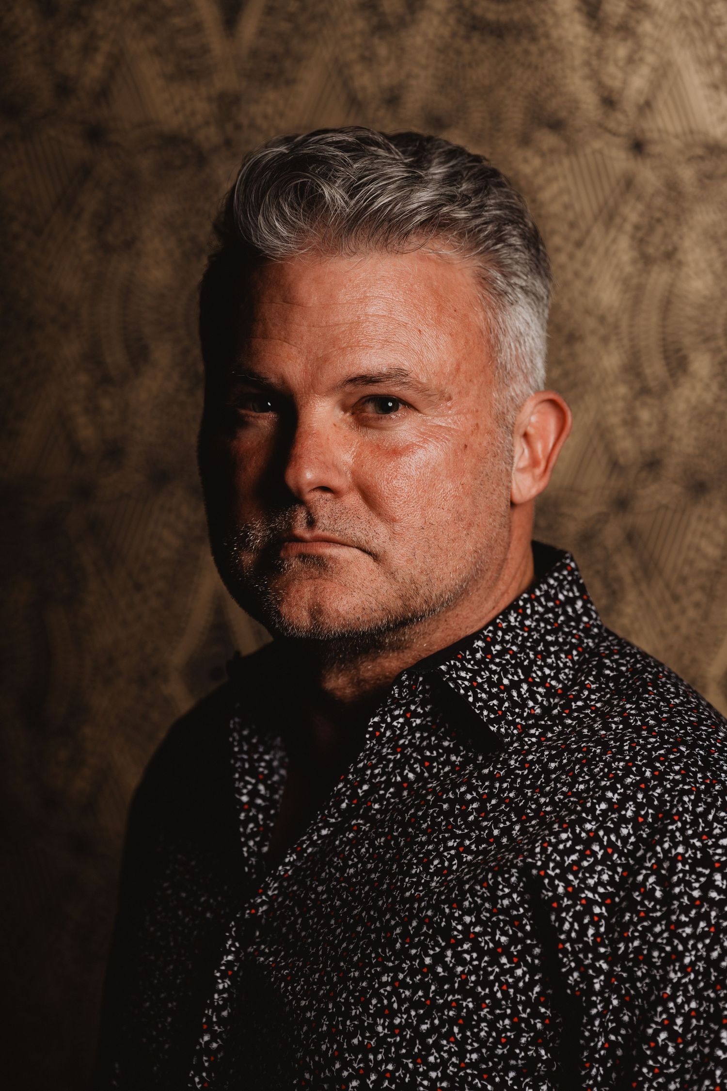Dramatic studio portrait of a man in patterned shirt against textured backdrop.