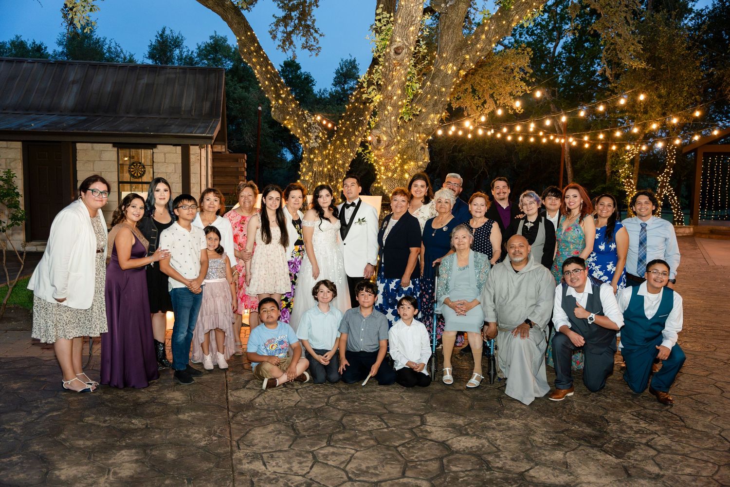 Groups of wedding guests gather outdoors under string lights during an evening celebration at a rustic venue setting.