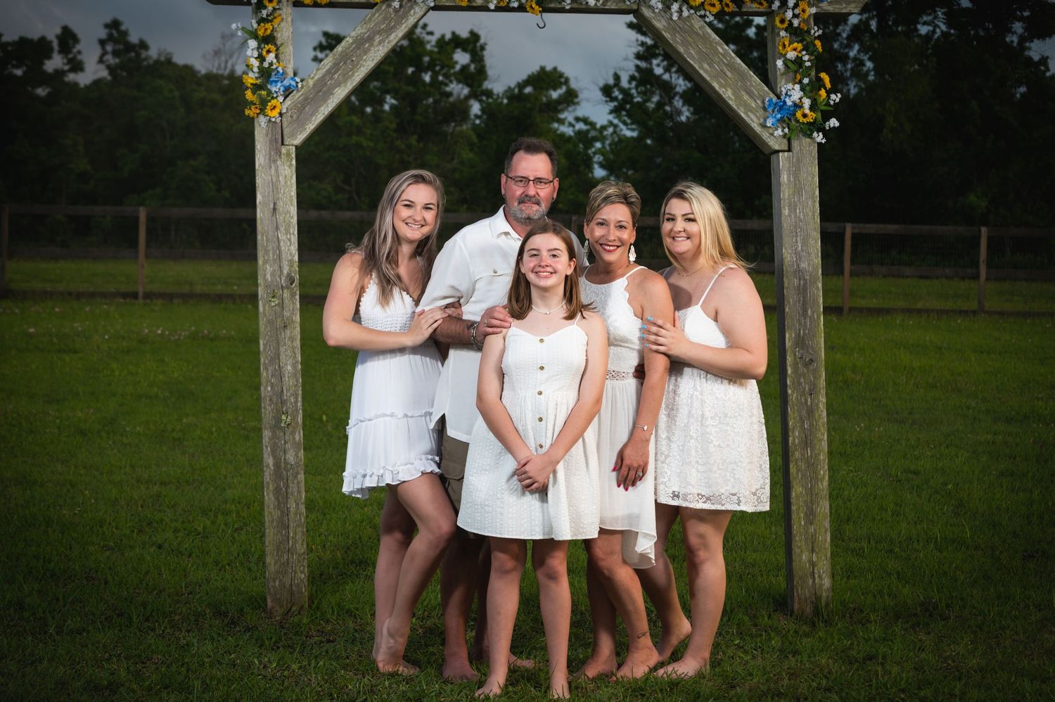 A family group stands barefoot under a wooden arch decorated with yellow flowers for a casual outdoor portrait.