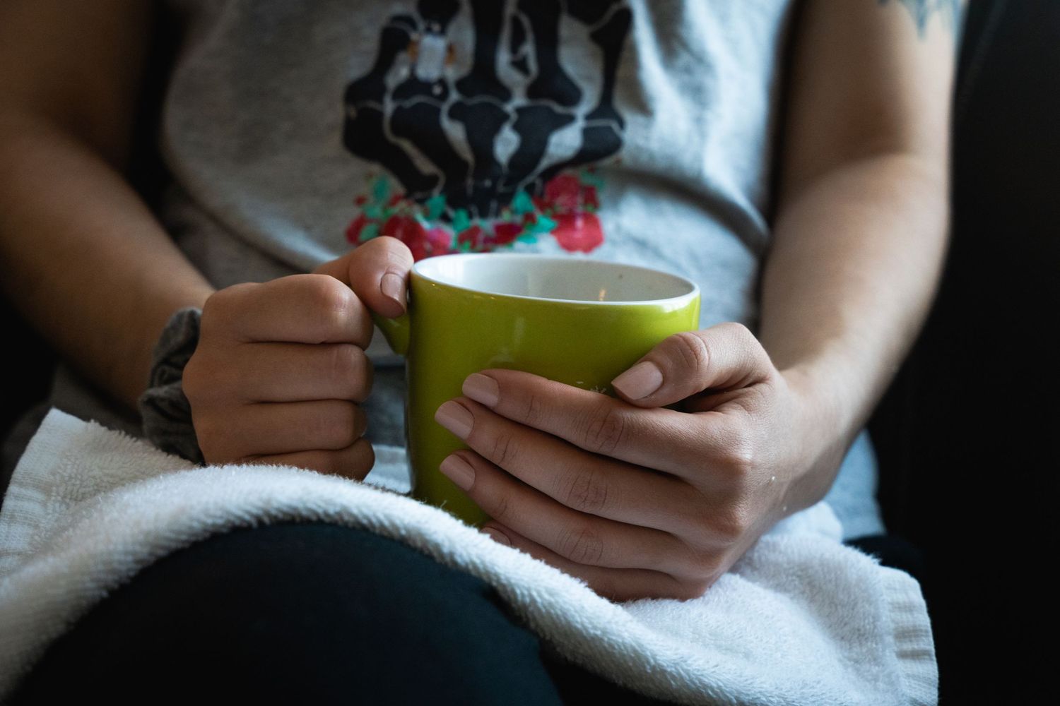 Hands holding a bright green mug while relaxing under a cozy blanket.