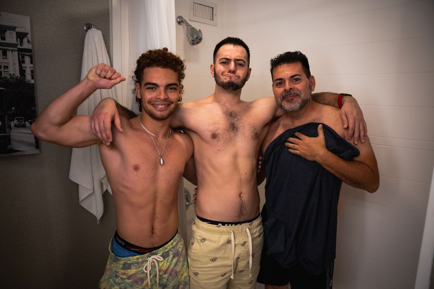 Three friends posing together shirtless in bathroom setting.