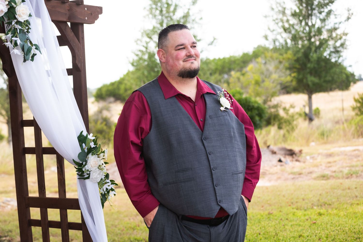 A person in formal attire stands under a wooden arch decorated with white fabric.