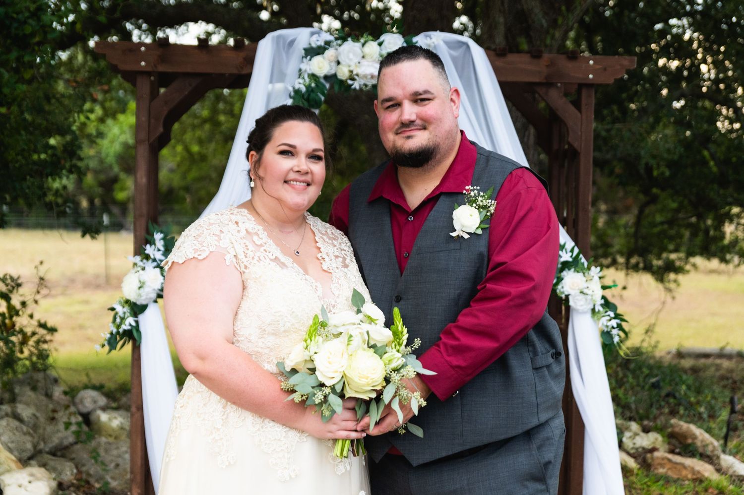 Happy couple poses under wooden wedding arch decorated with white flowers and drapery.
