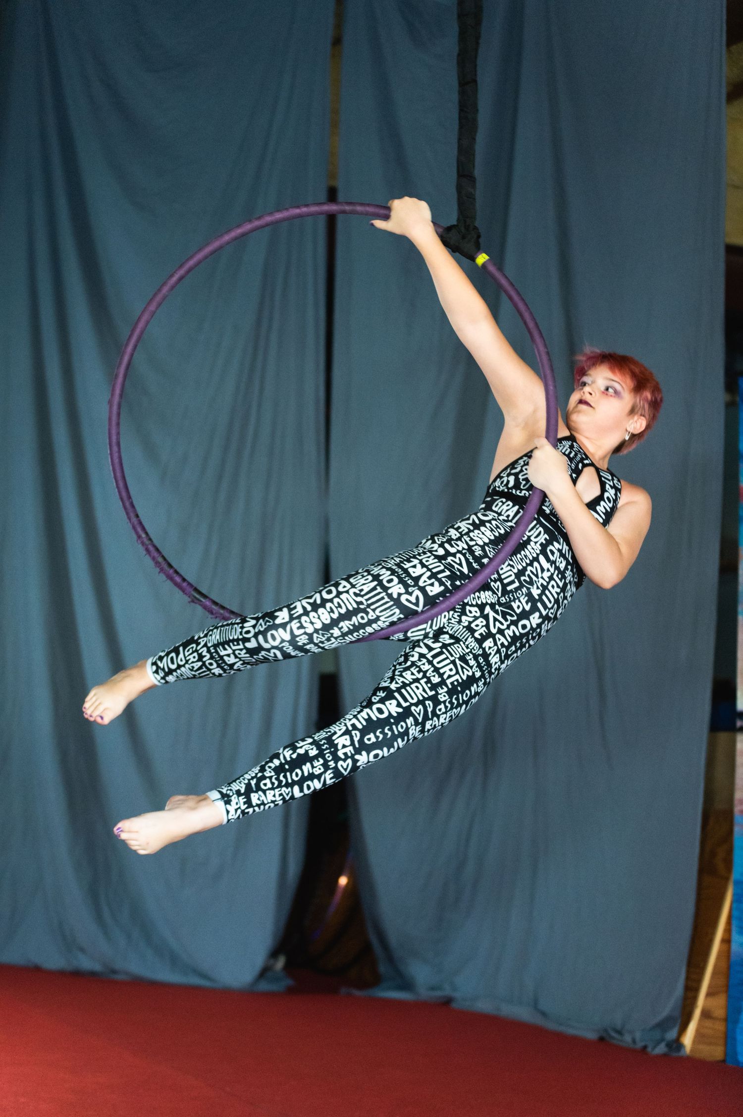 Performer executes graceful aerial hoop pose in printed jumpsuit against blue backdrop.