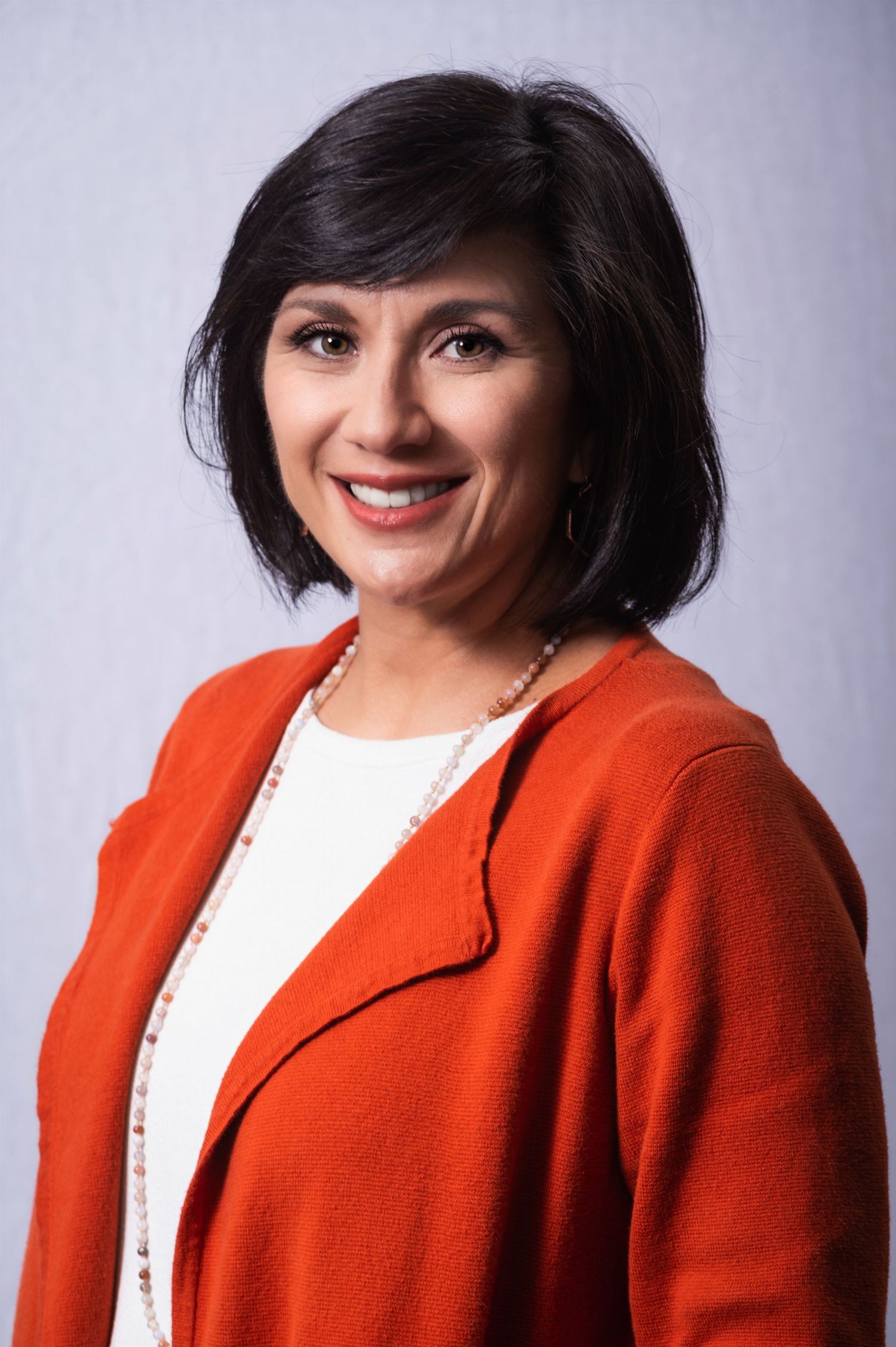 Professional portrait of someone wearing a bright orange cardigan and white top with a beaded necklace.