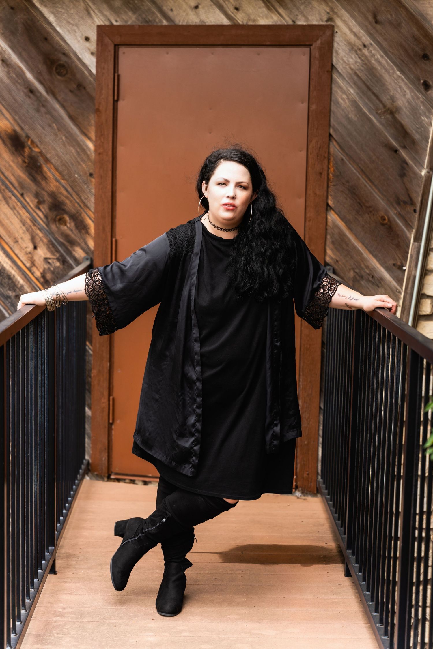 Figure in flowing black dress poses on wooden walkway against orange door.