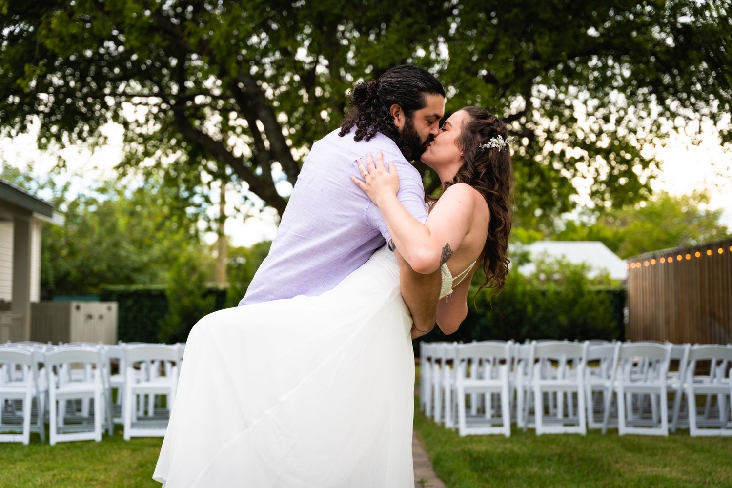A romantic wedding couple sharing a kiss in a backyard setting with white chairs.