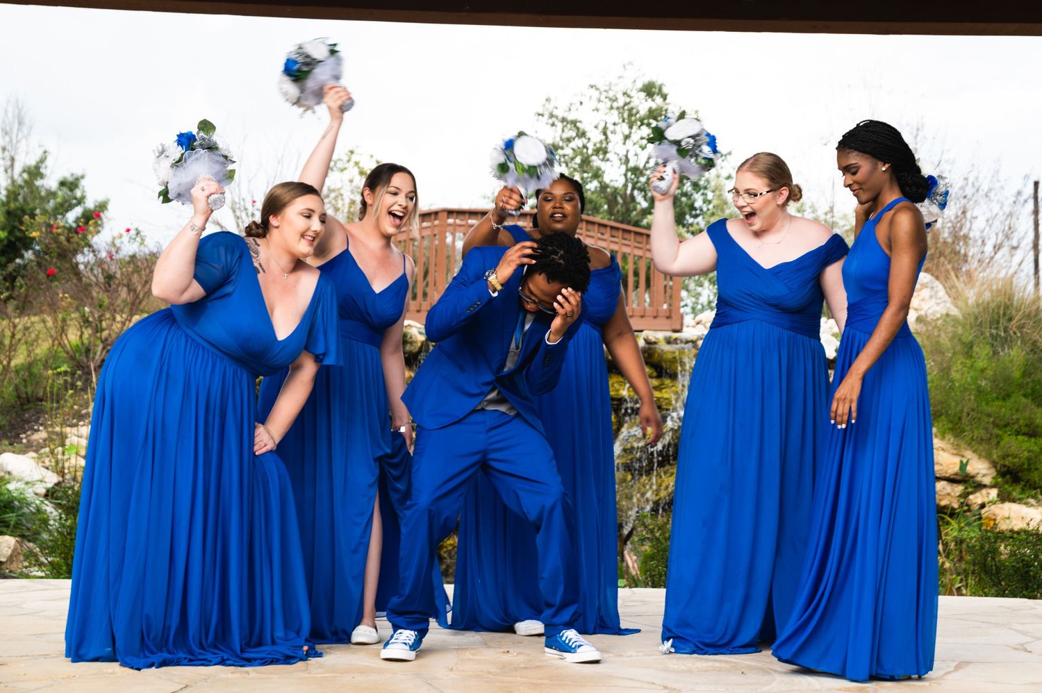 Wedding party members in royal blue dresses celebrate joyfully with bouquets outdoors.