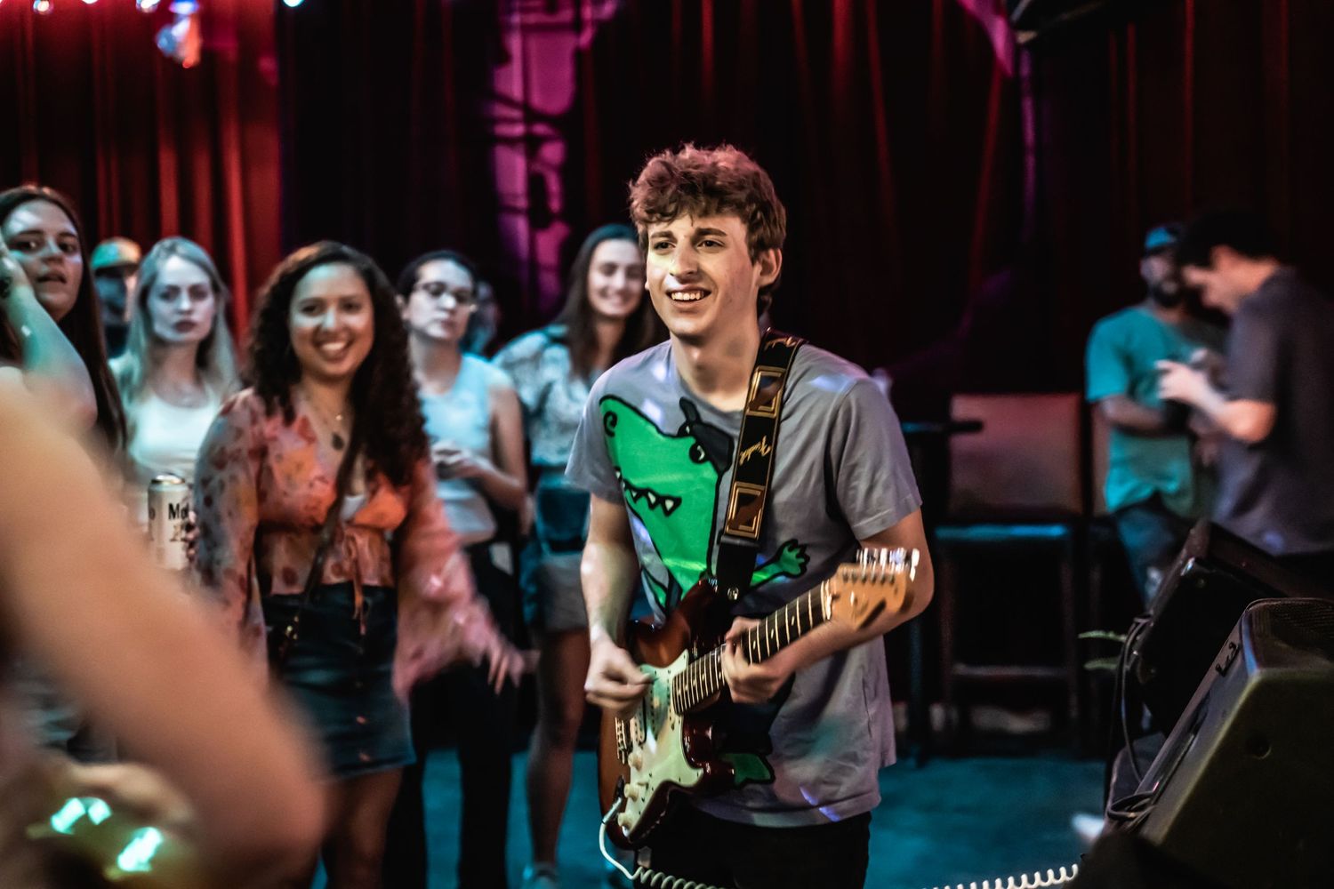 A guitarist performs on stage at a music venue with colorful lighting and crowd in background.