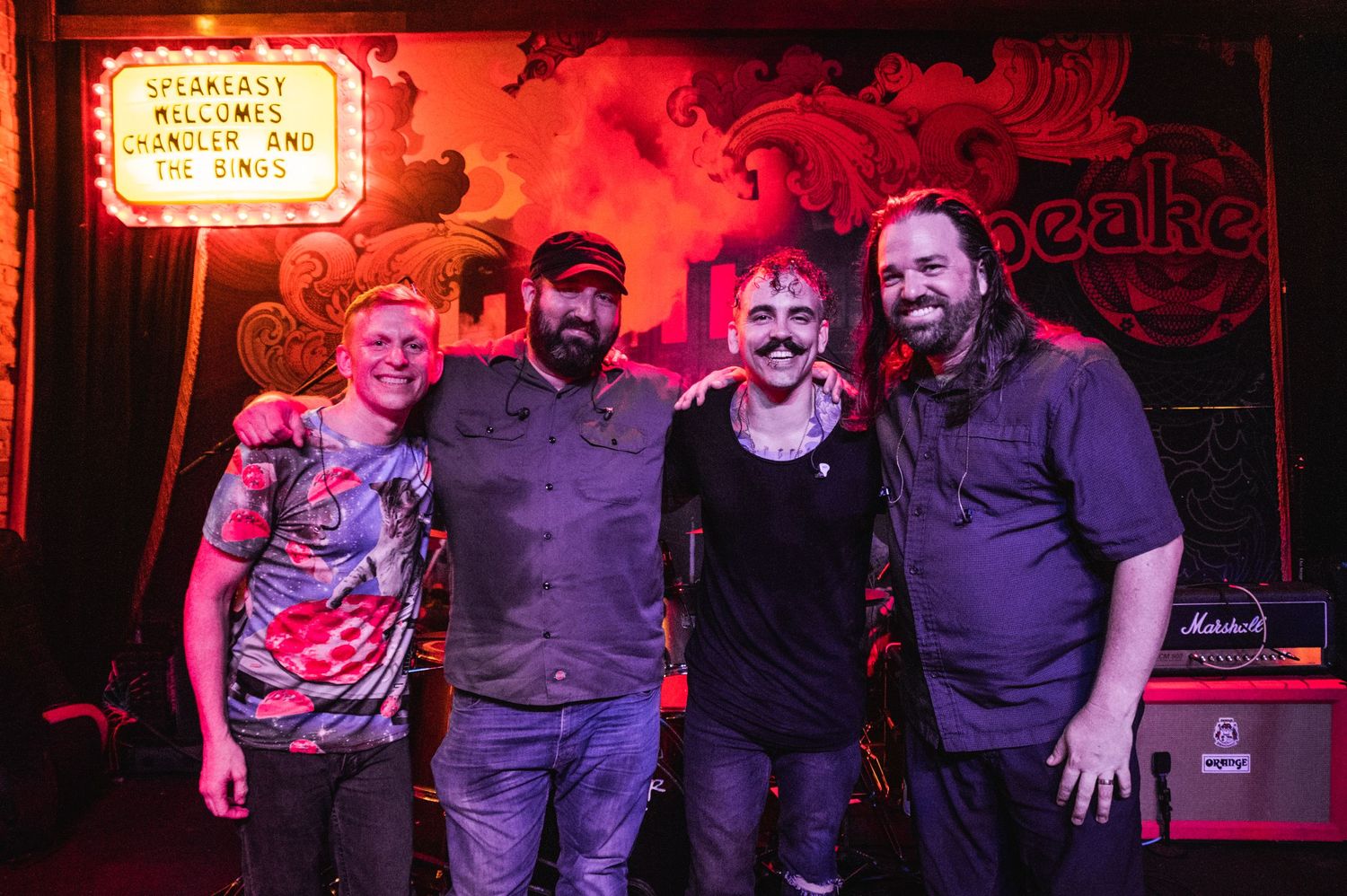 Four musicians pose together under red lighting at a live music venue.