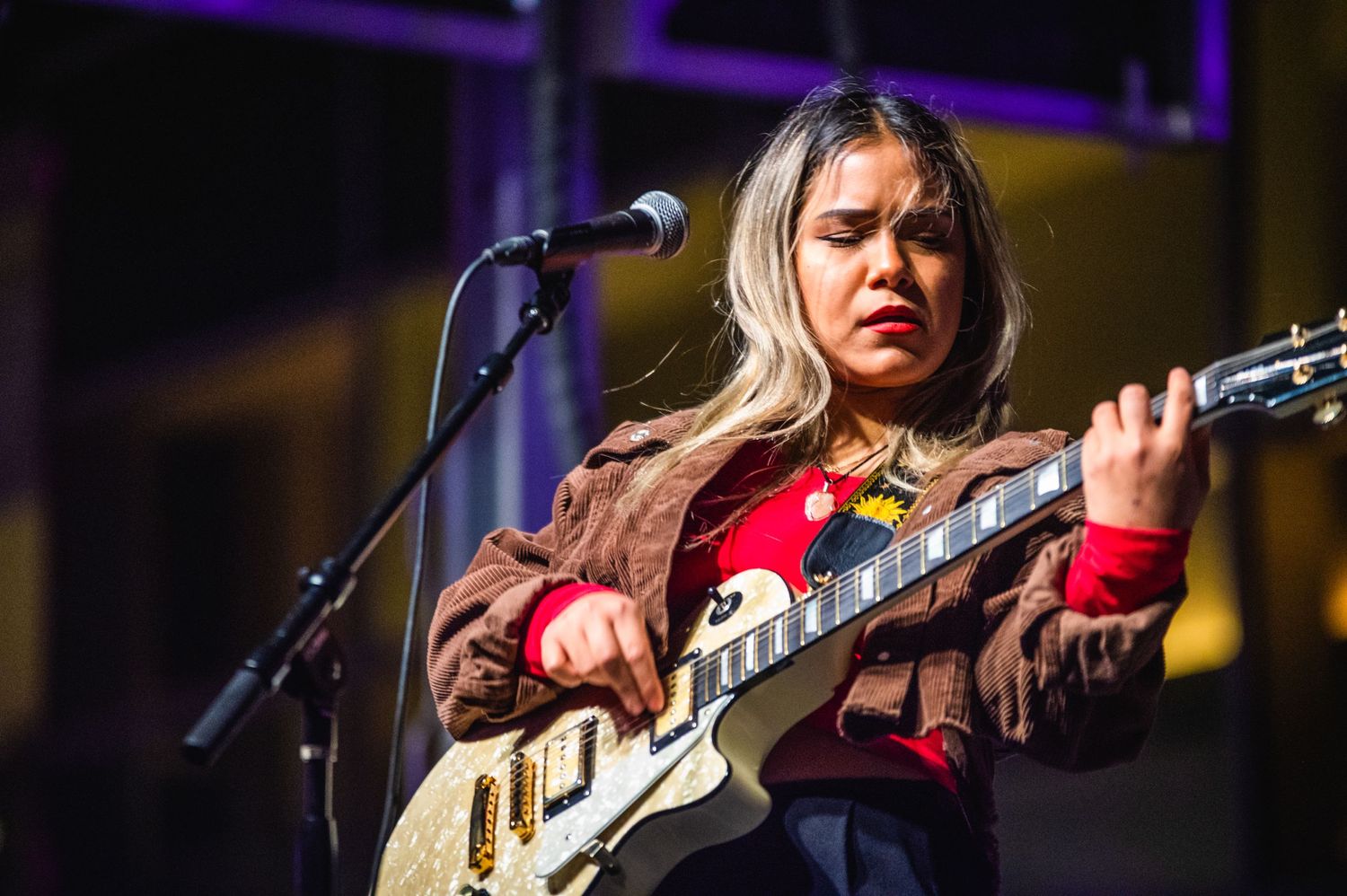 Guitarist in red shirt plays white electric guitar while singing into microphone on stage.