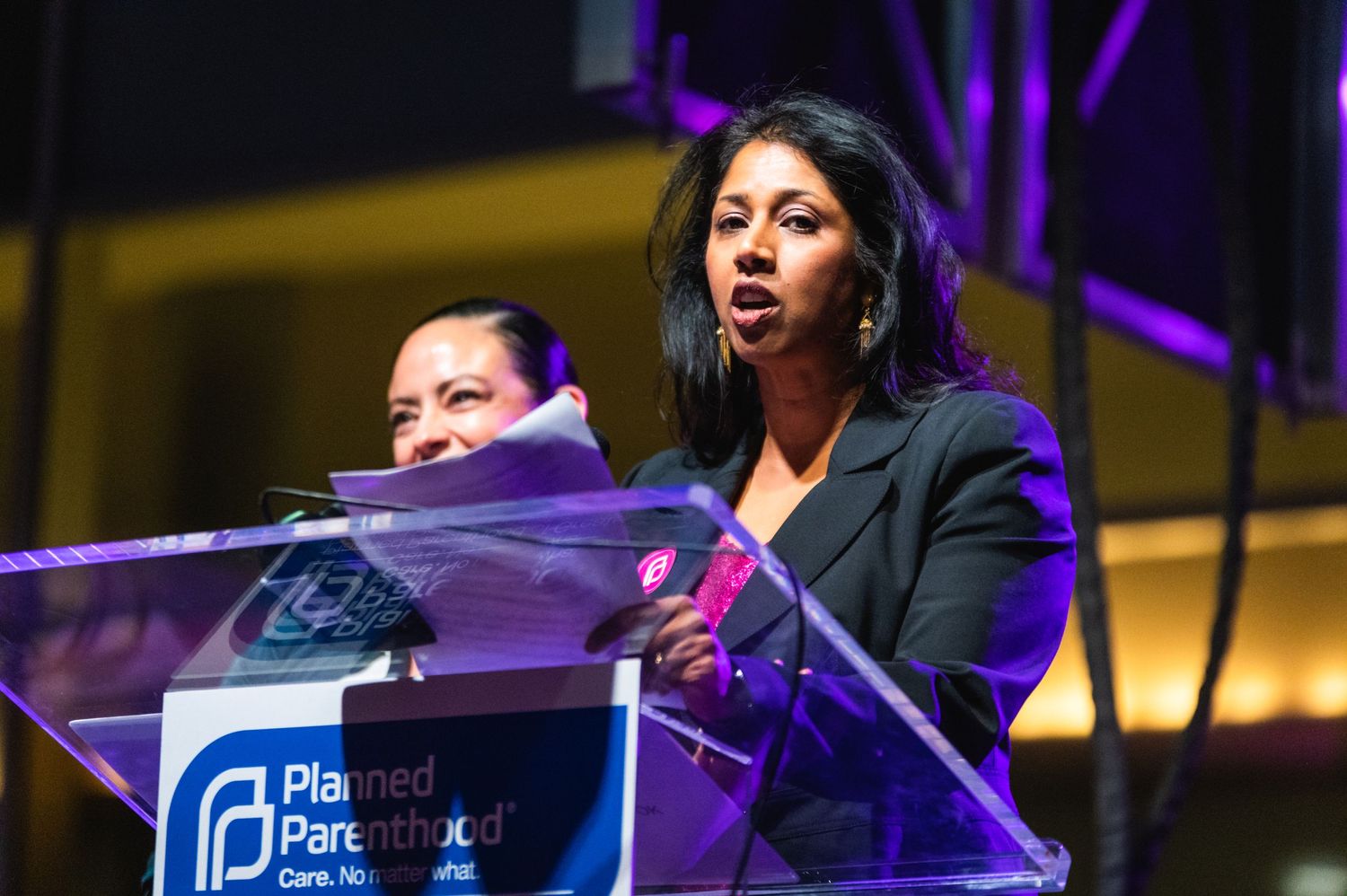 Presenter in dark blazer speaks at illuminated podium during organizational event.