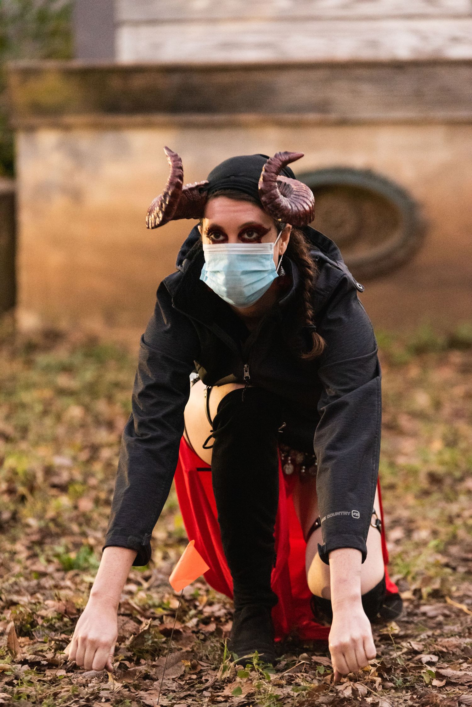 A person in a dark coat and face mask crouches low in fallen autumn leaves wearing butterfly decorations.
