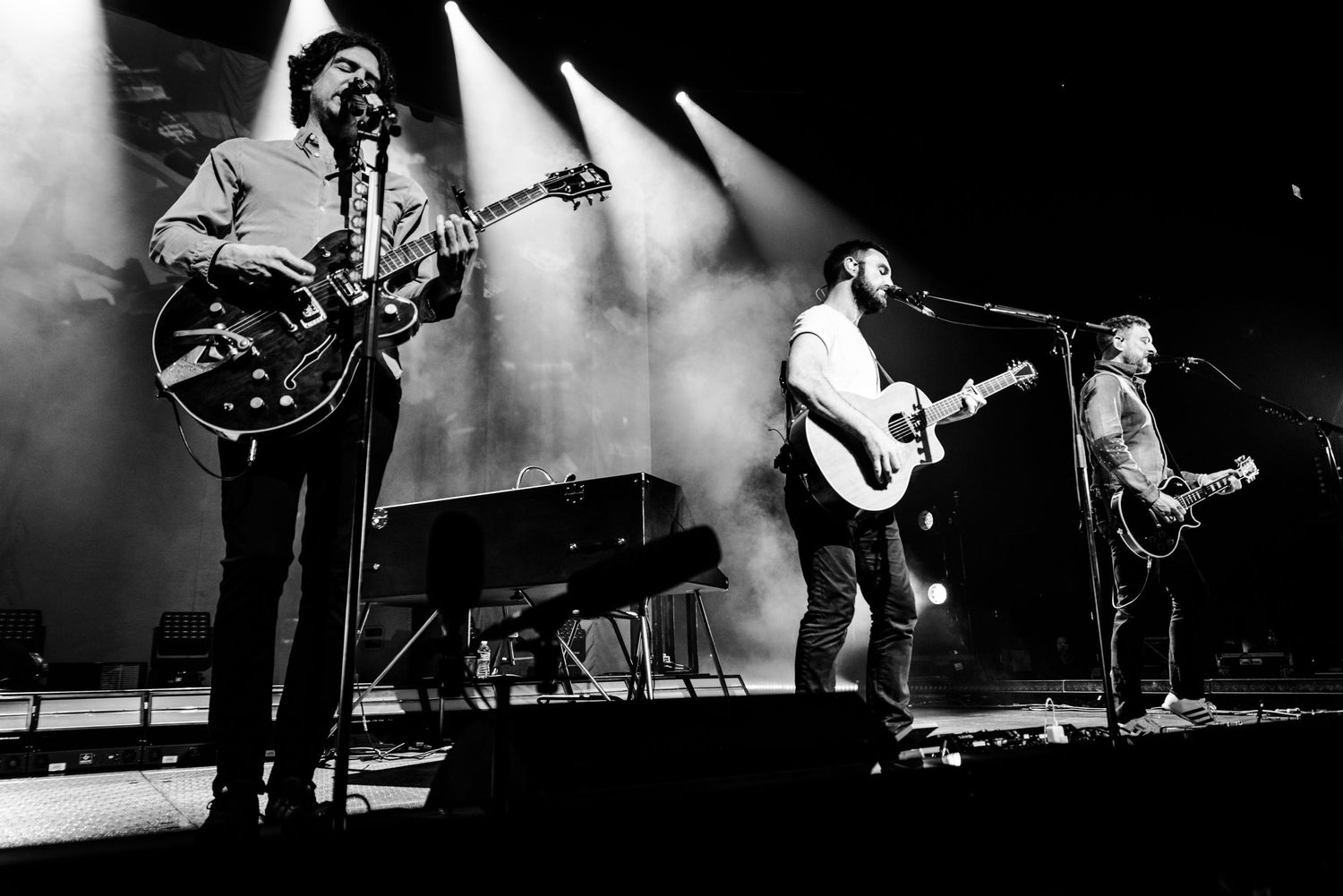 Black and white wide shot of three musicians performing with guitars on concert stage.