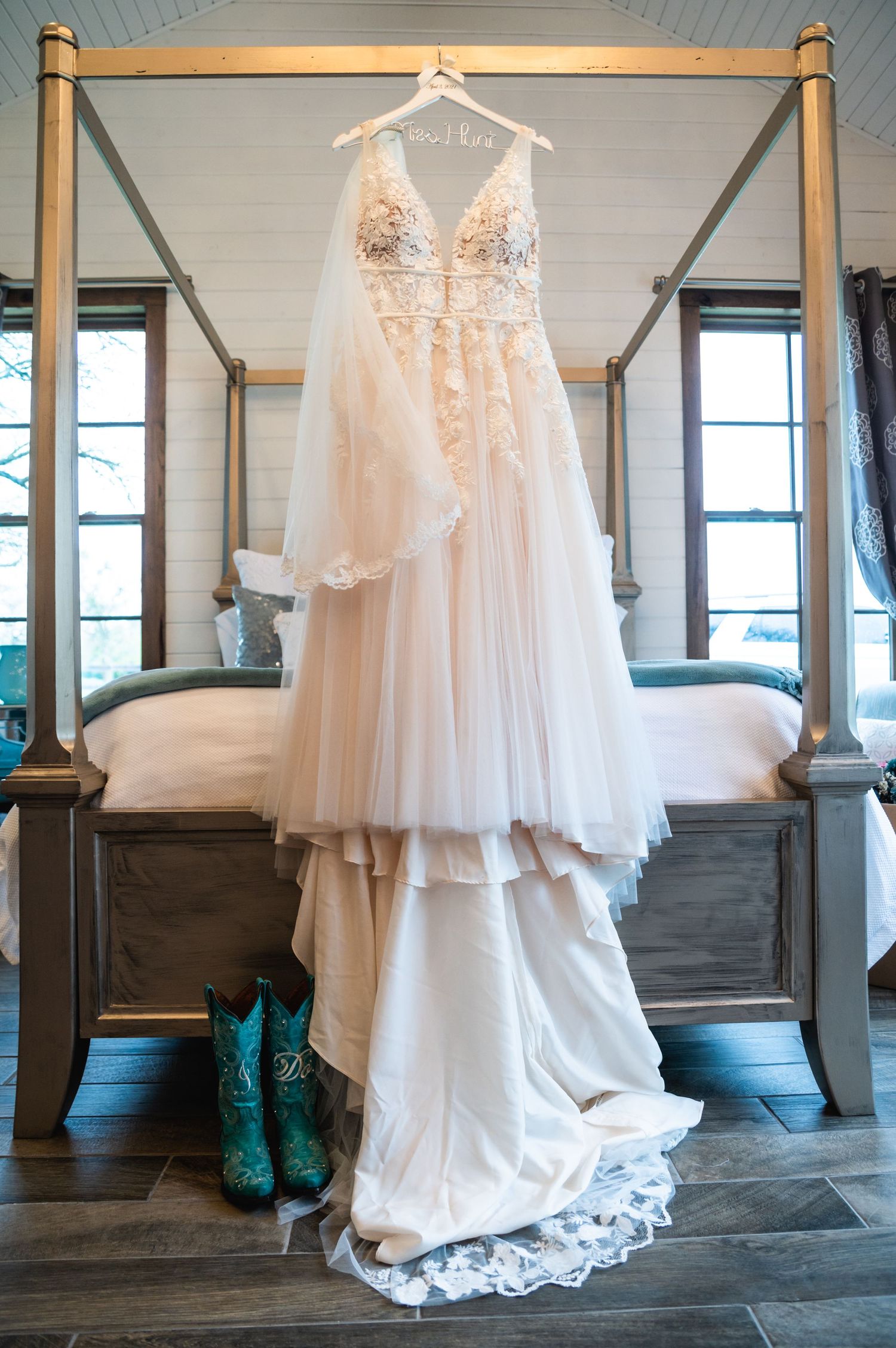 Elegant white wedding dress with lace details hanging on a rustic four-poster bed with turquoise cowboy boots below.