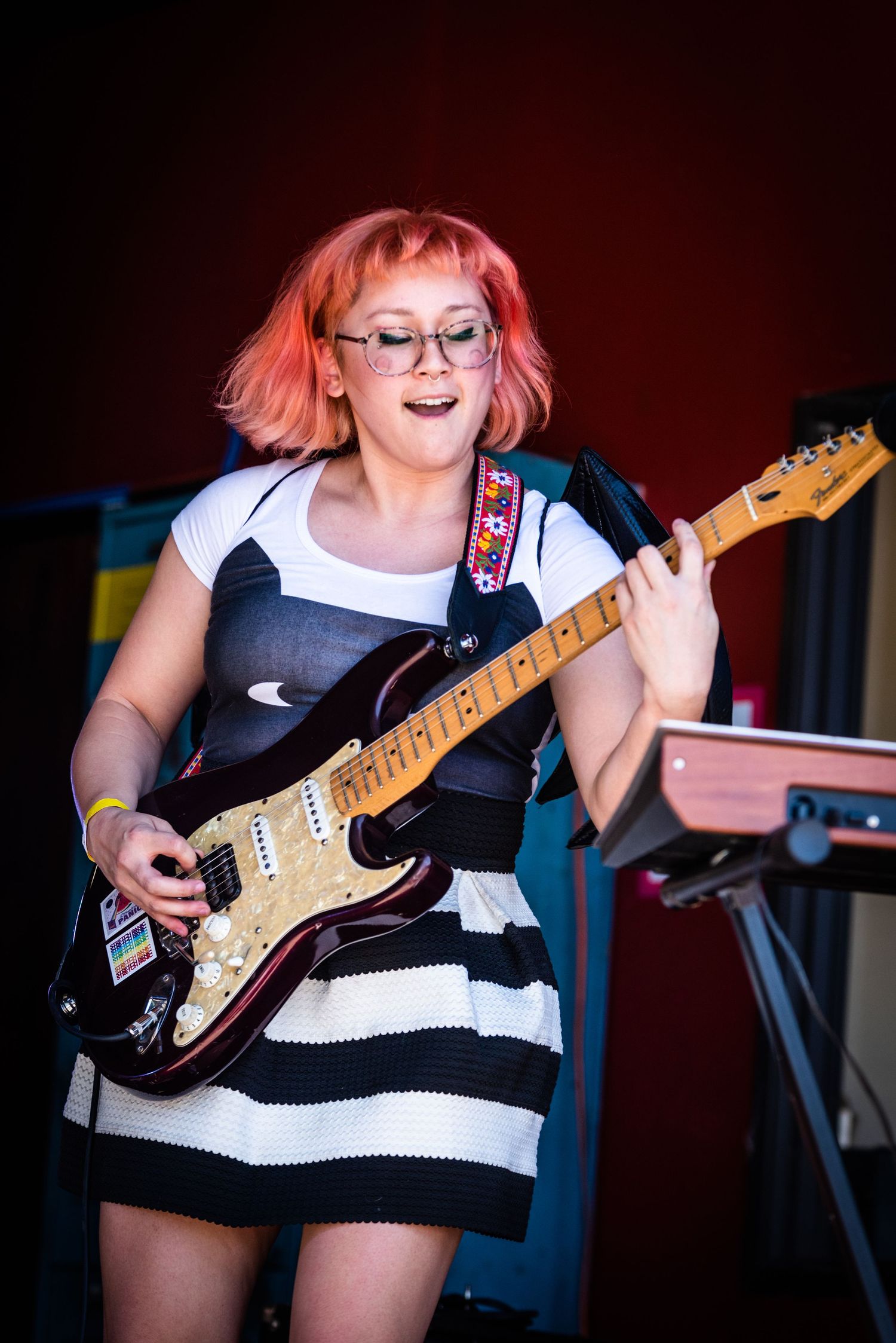 Musician in black and white striped dress plays electric guitar on stage with red lighting effects.