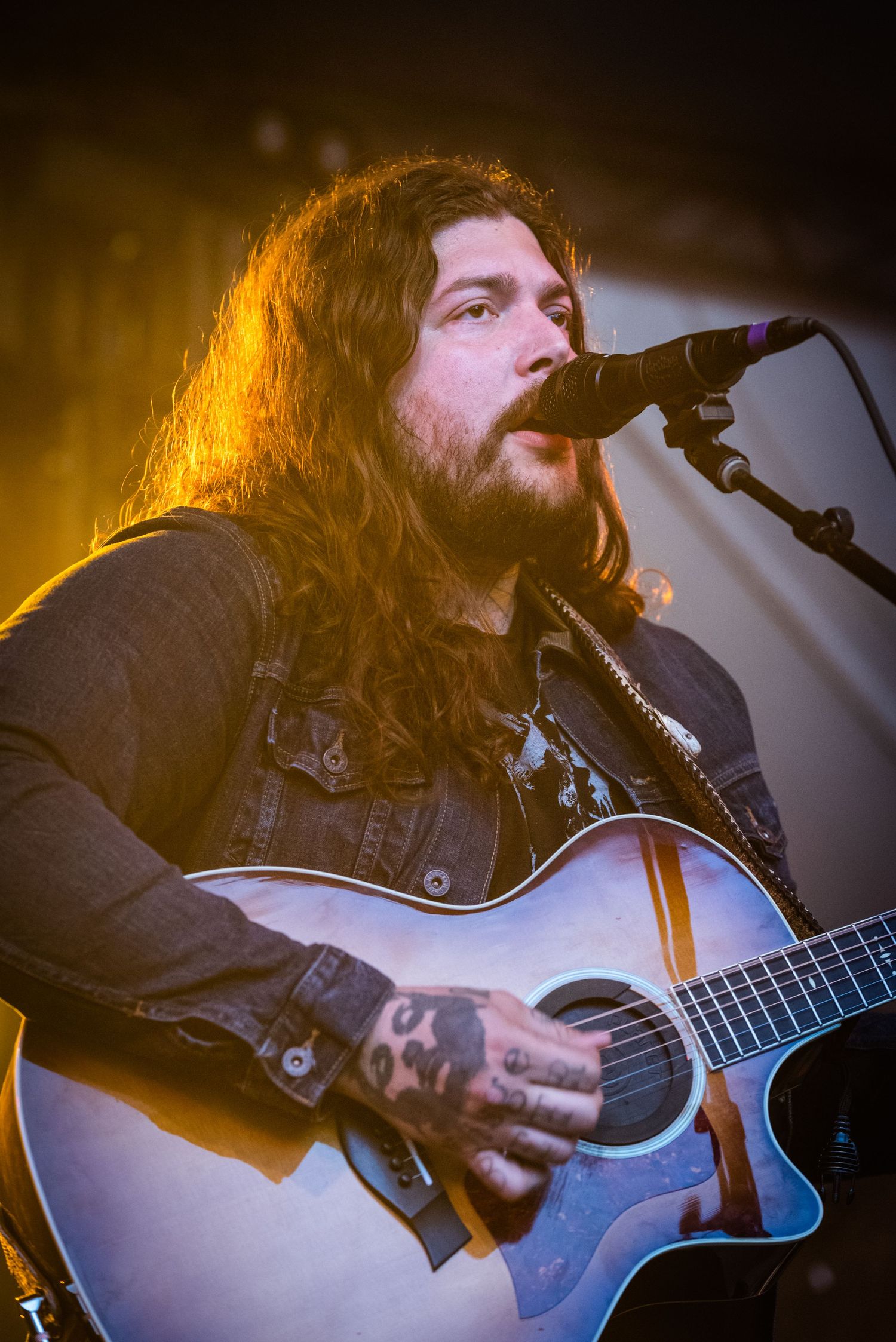Folk musician with flowing hair performs on acoustic guitar under warm stage lights.