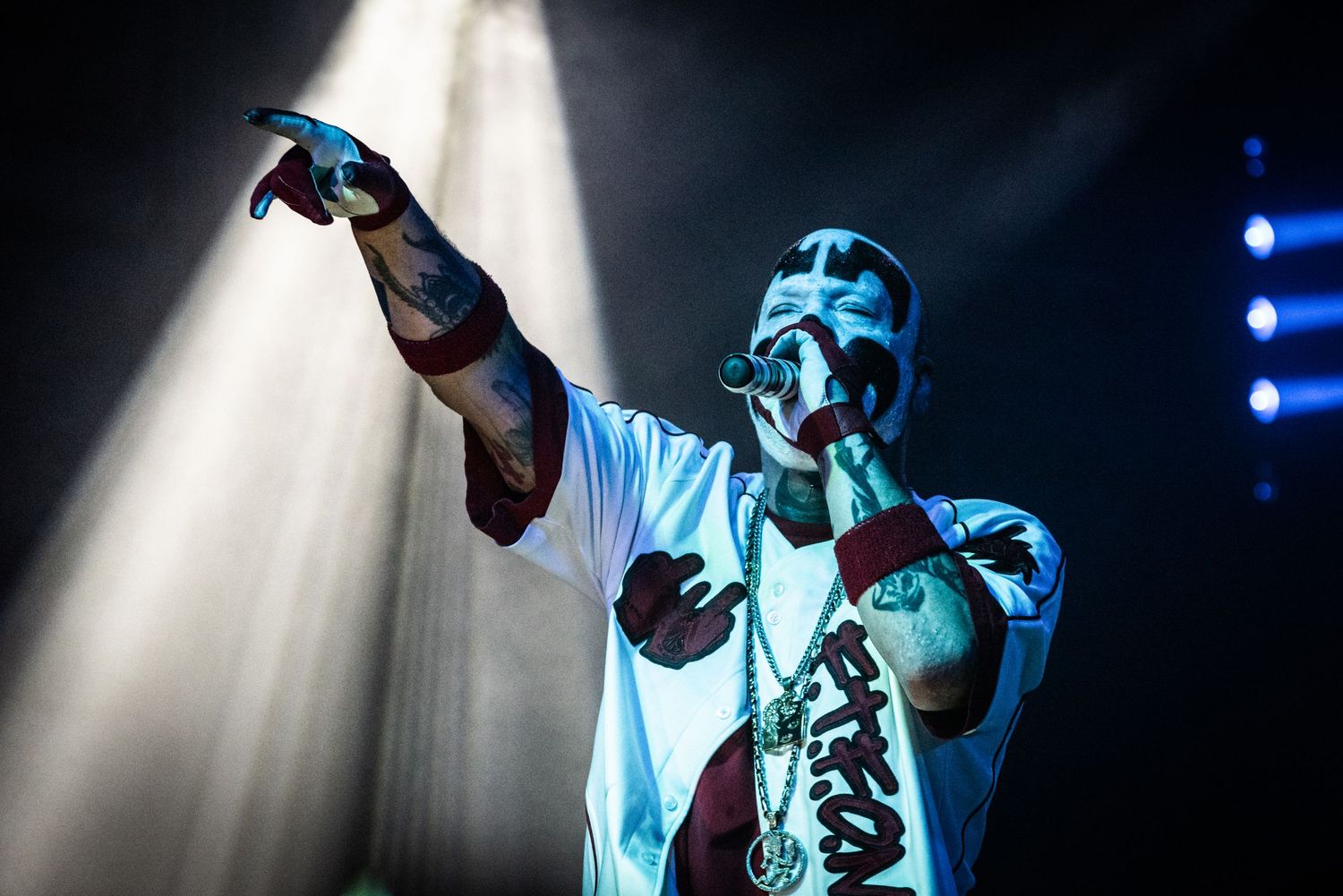 Dramatic concert lighting illuminates performer in white costume with outstretched arm.